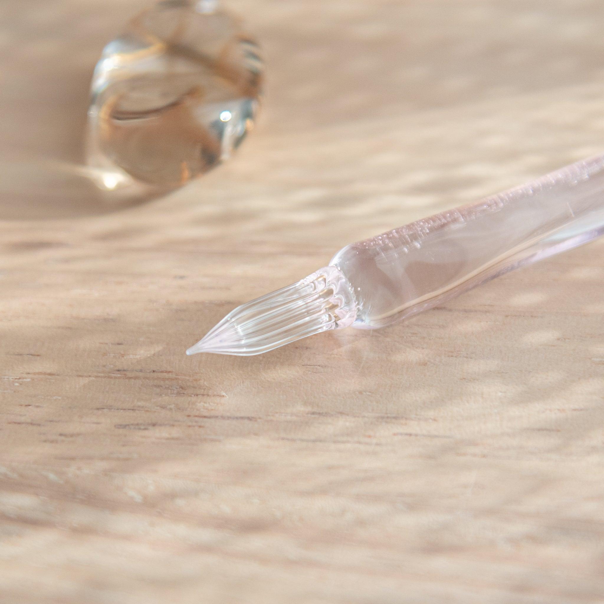 Close-up of a glass dip pen’s nib with subtle pink tint, resting on a light wooden surface, next to a small, rounded glass pen rest.