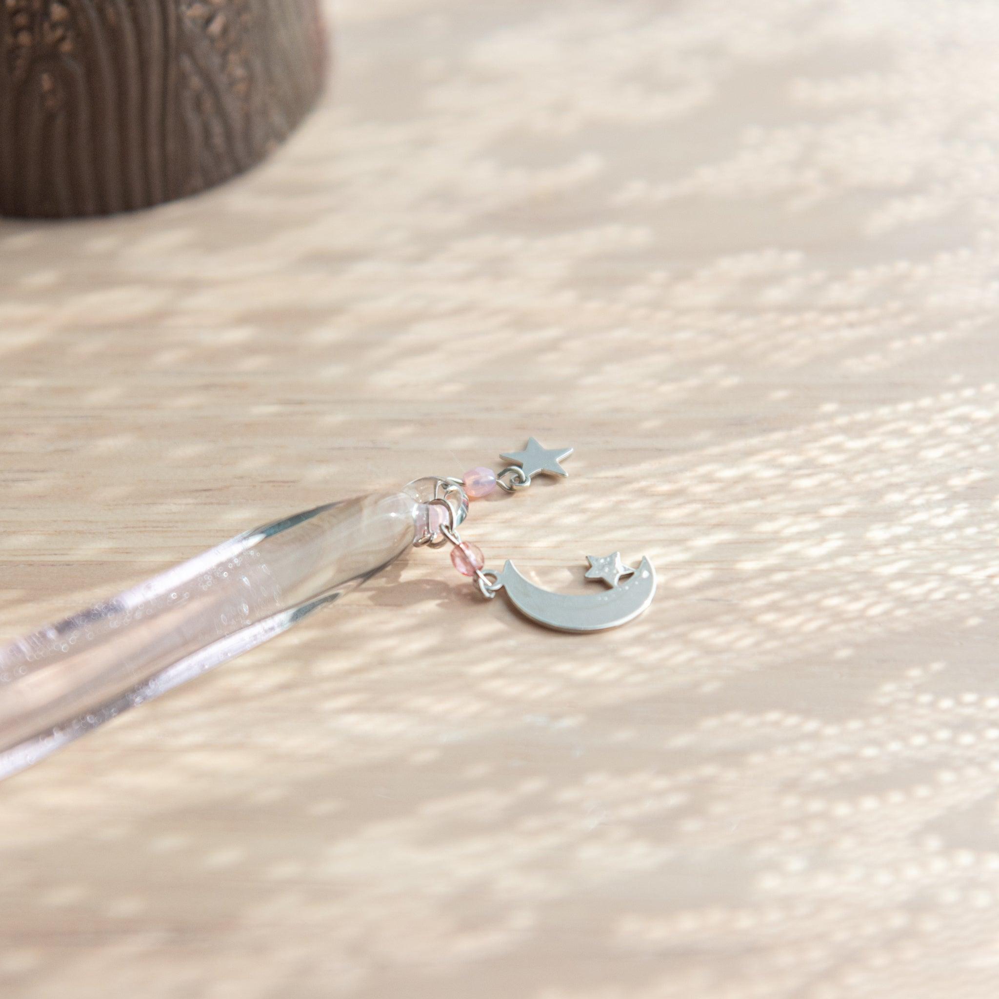 Moon and star charm with pink bead detail attached to a glass dip pen, lying on a light wooden surface with soft, dappled light.