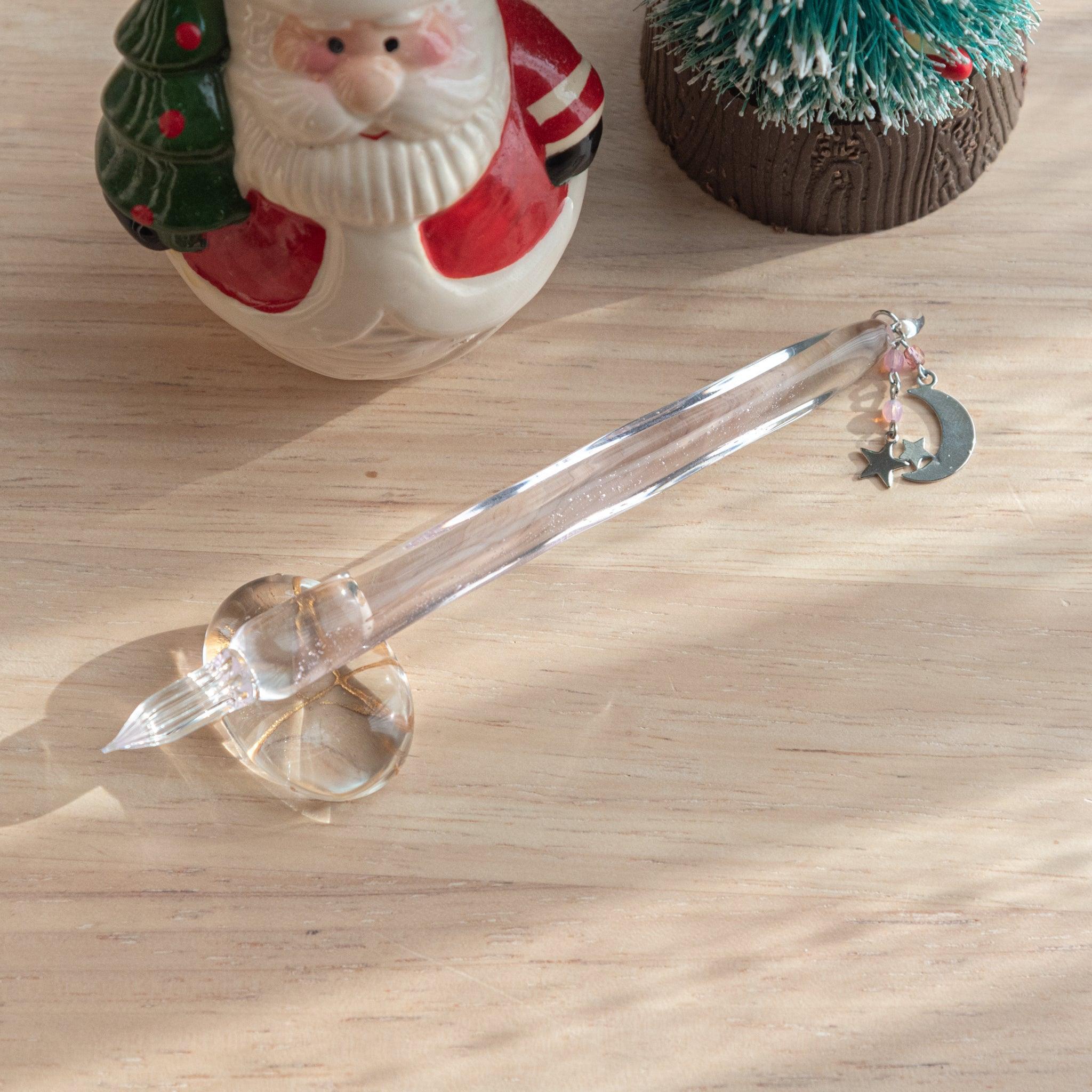 Glass dip pen with a moon and star charm, placed on a light wooden surface near a Santa figurine and a small tree decoration.