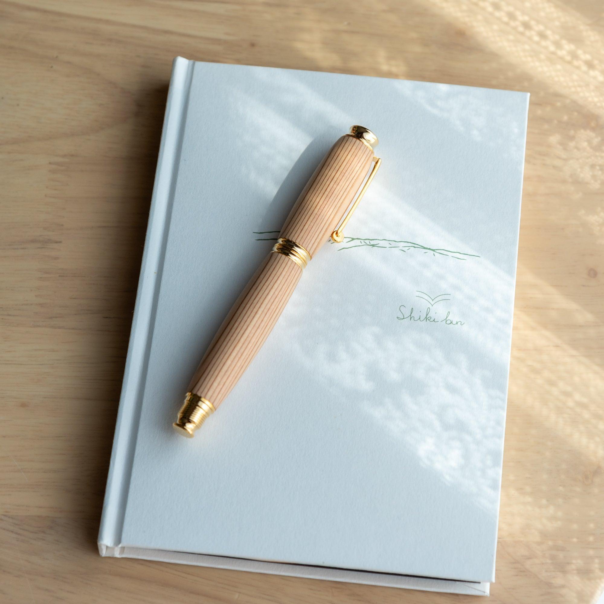 A Japanese Yakushima cedar wood fountain pen with gold details, elegantly resting on a white notebook to highlight its natural and refined design.