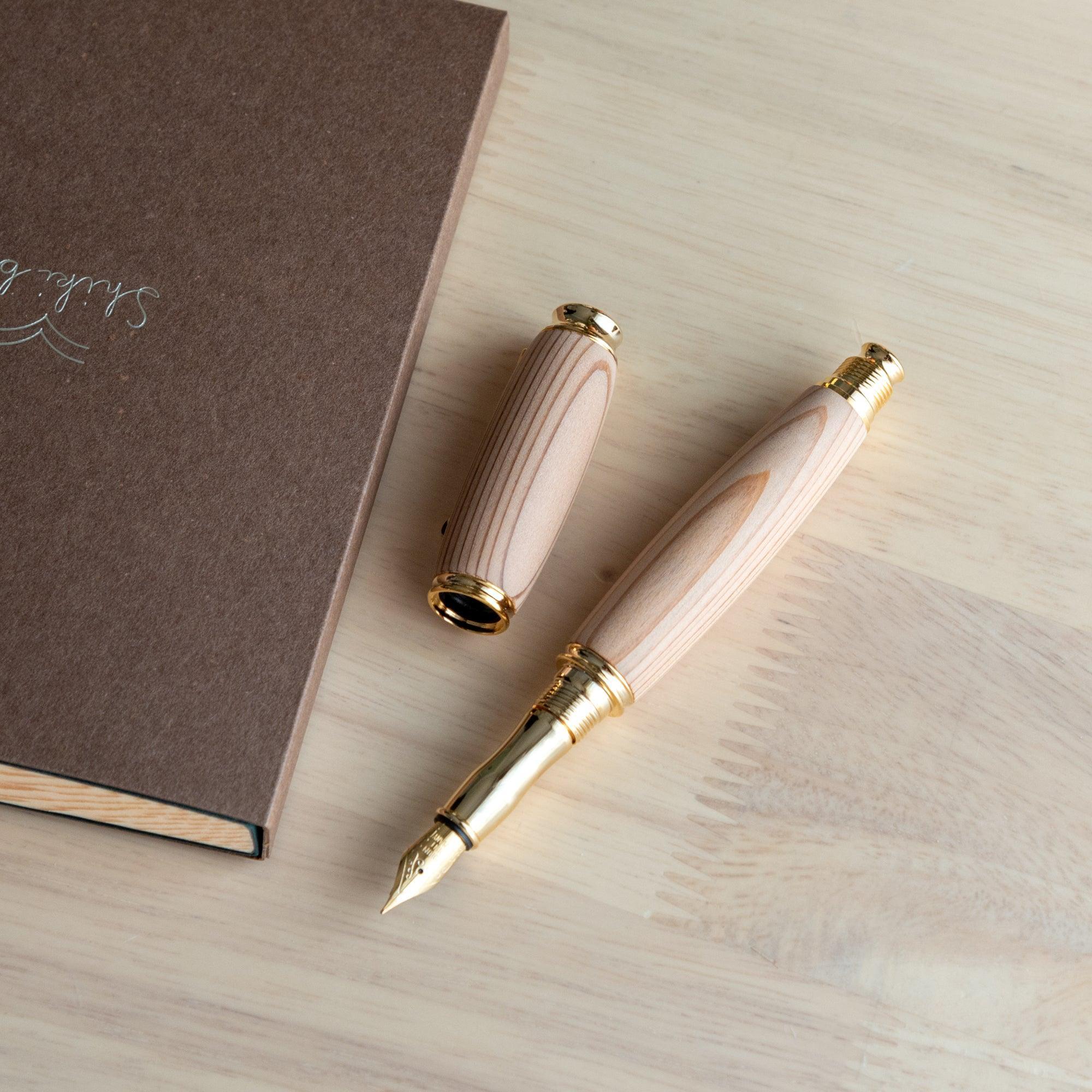 A Japanese fountain pen made of Yakushima cedar wood, featuring a gold nib and detachable cap, displayed open next to a brown notebook on a wooden desk.