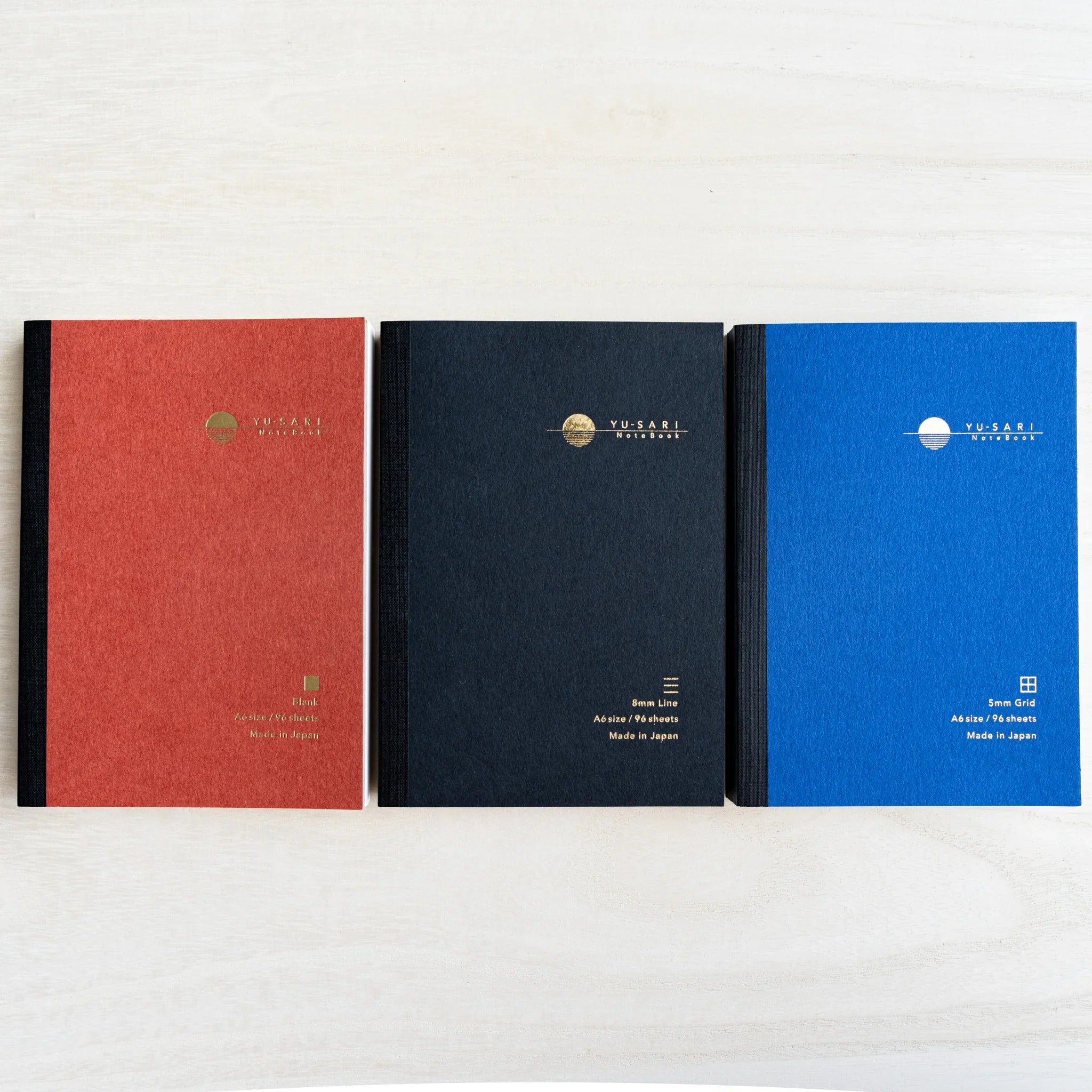 Three A6 YU-SARI notebooks lying side by side on wood, dark red Blank, black 8mm Line, and blue 5mm Grid with gold logos