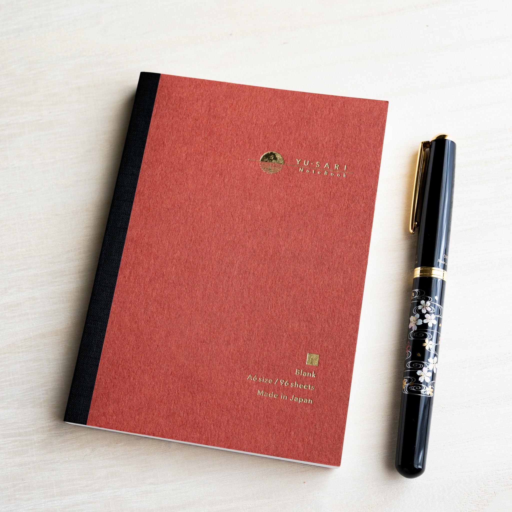 Dark red A6 YU-SARI blank notebook on wood beside a black lacquer maki-e fountain pen, gold Blank, A6 size, 96 sheets text