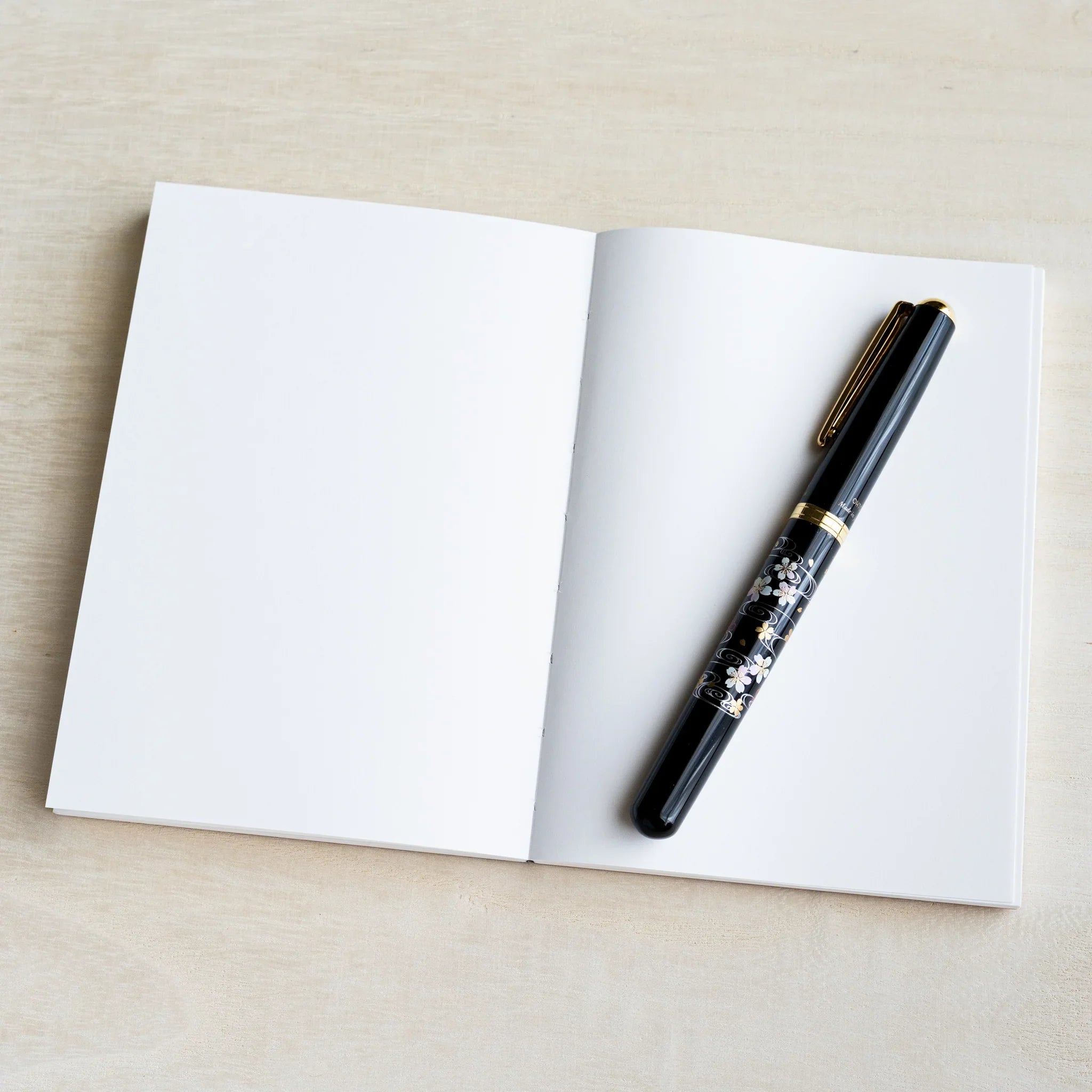 Open blank A6 YU-SARI notebook on wood showing two white pages with a black lacquer maki-e fountain pen on the right page