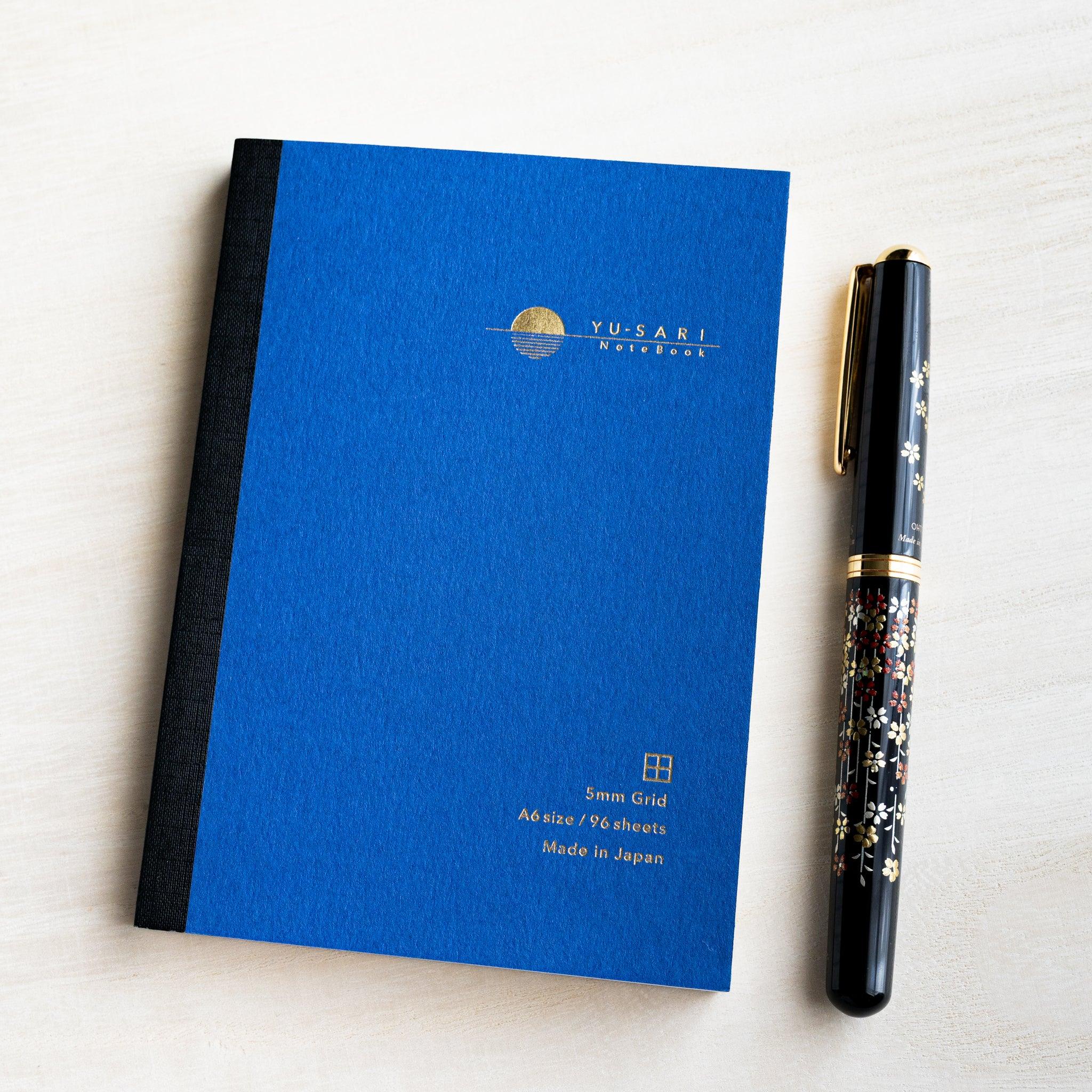 Blue A6 YU-SARI grid notebook on wood beside a black lacquer maki-e fountain pen, gold 5mm Grid, A6 size, 96 sheets text