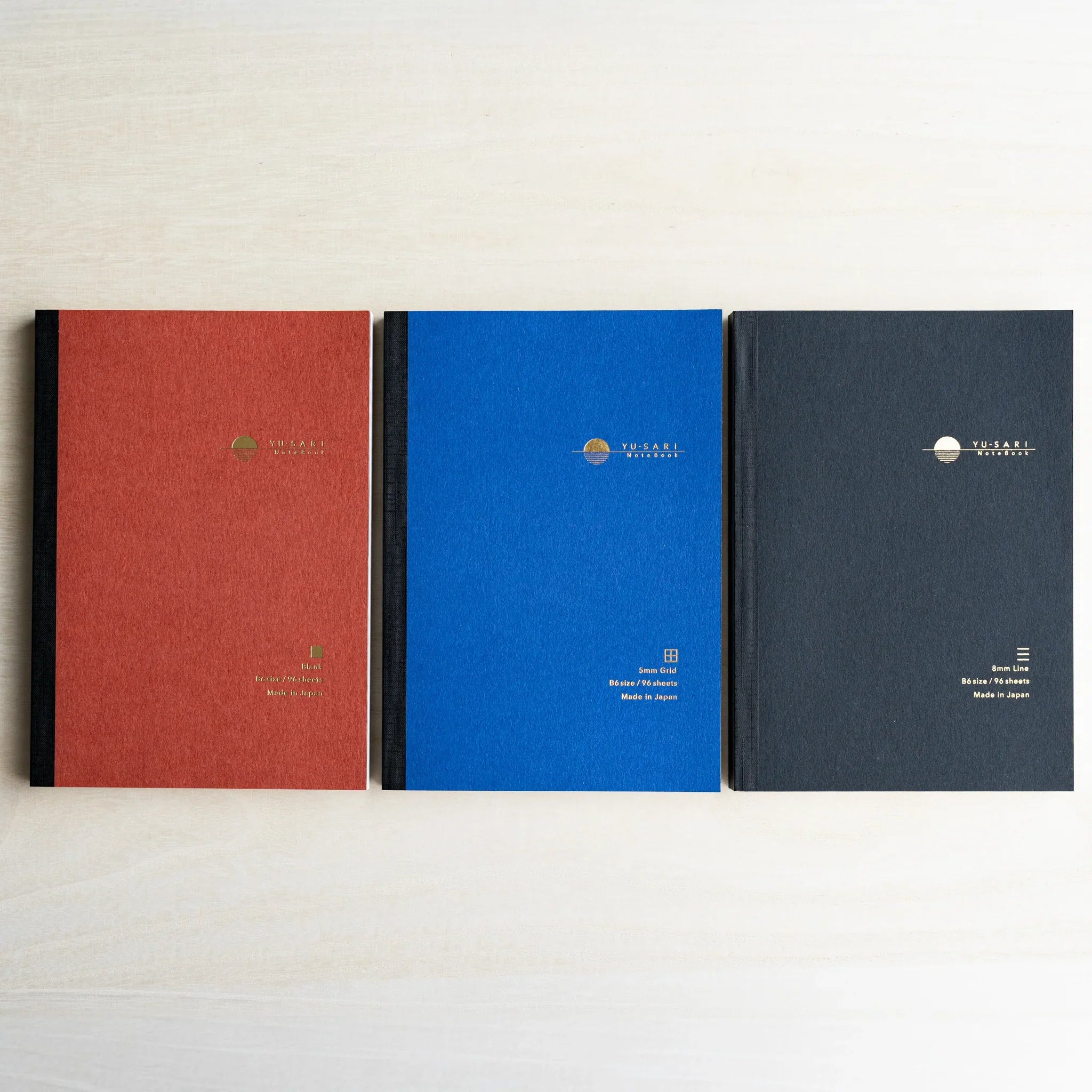 Three B6 YU-SARI notebooks lying side by side on wood, dark red Blank, blue 5mm Grid, and black 8mm Line with gold logos