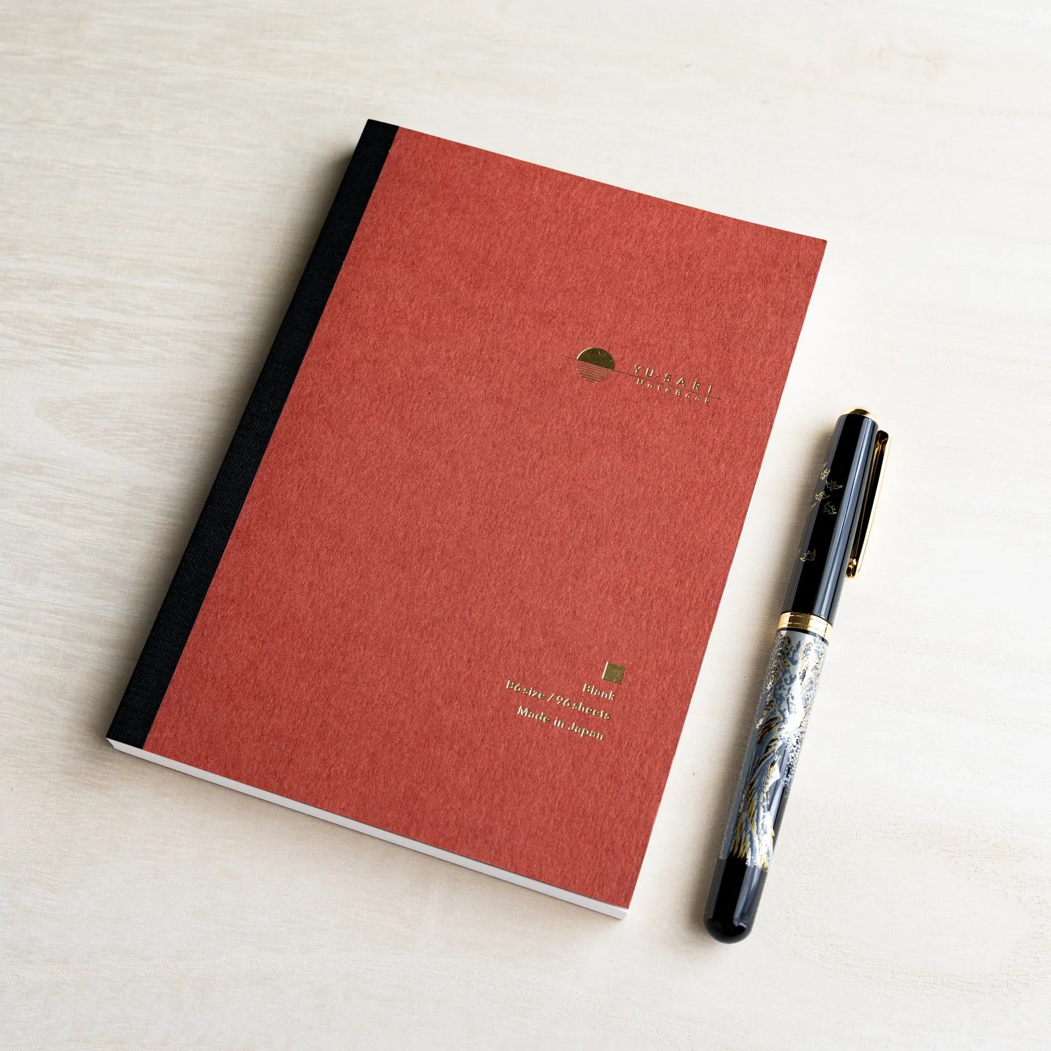 Dark red B6 YU-SARI blank notebook on wood beside a black lacquer maki-e fountain pen, gold Blank, B6 size, 96 sheets text