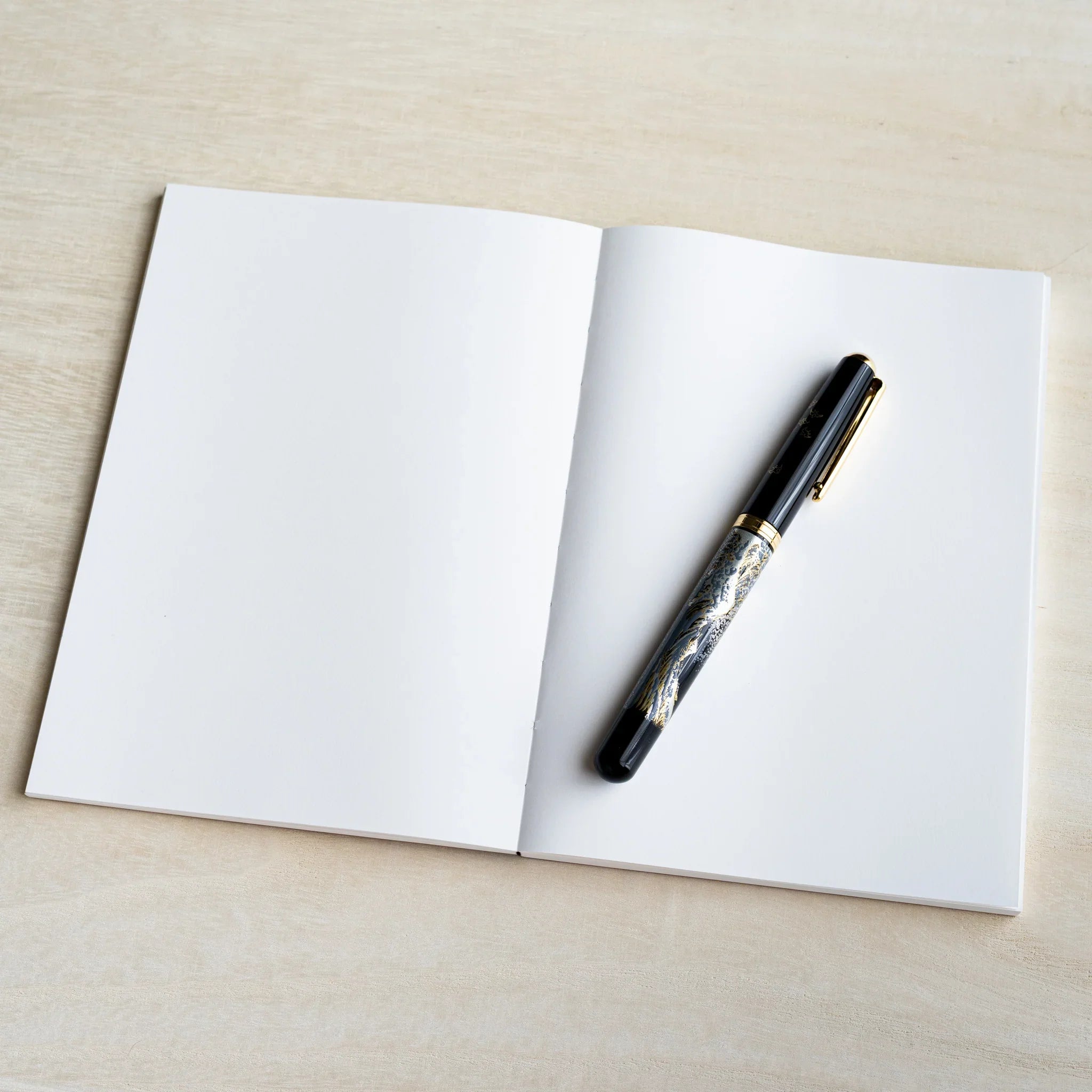 Open blank B6 YU-SARI notebook on wood showing two white pages with a black lacquer maki-e fountain pen on the right page