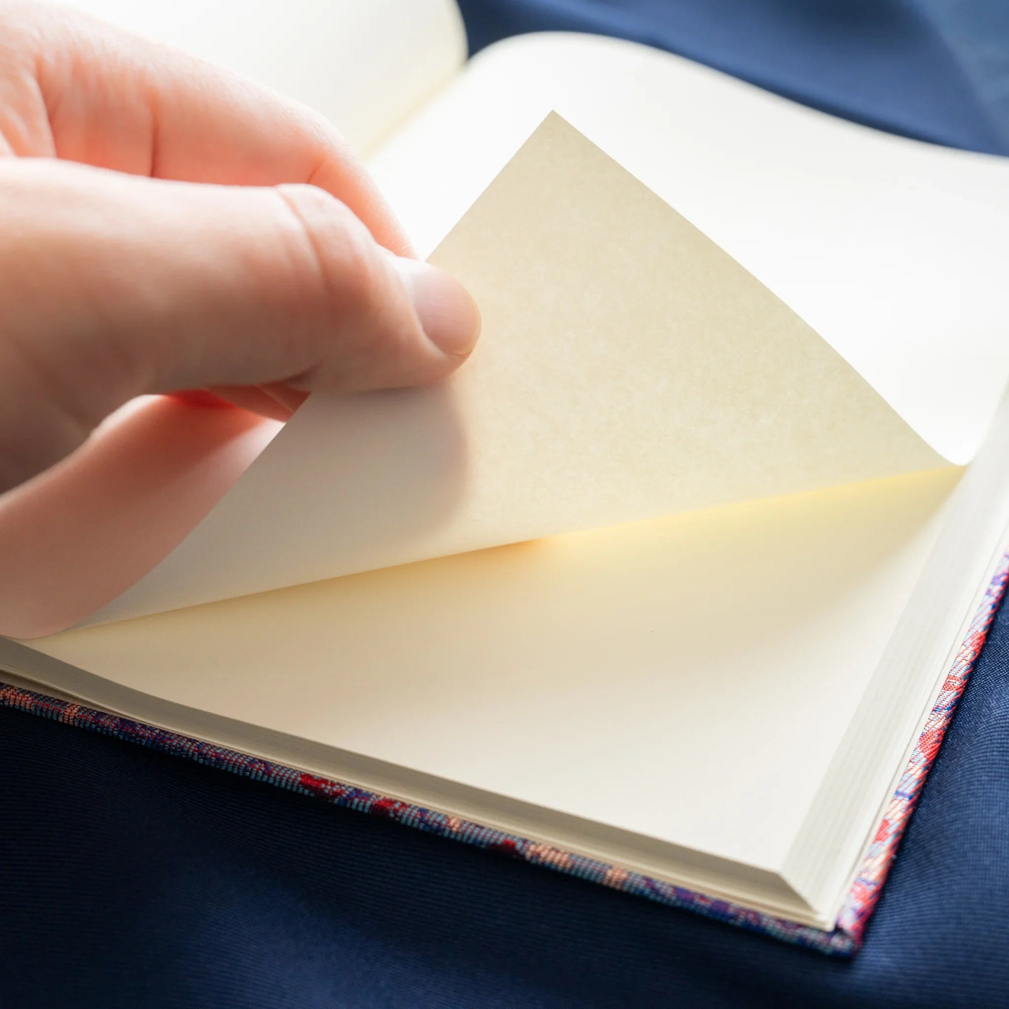 Hand turning ivory pages of open Yusoku Hanabishi Nishijin silk notebook, silk binding visible at bottom, blue fabric