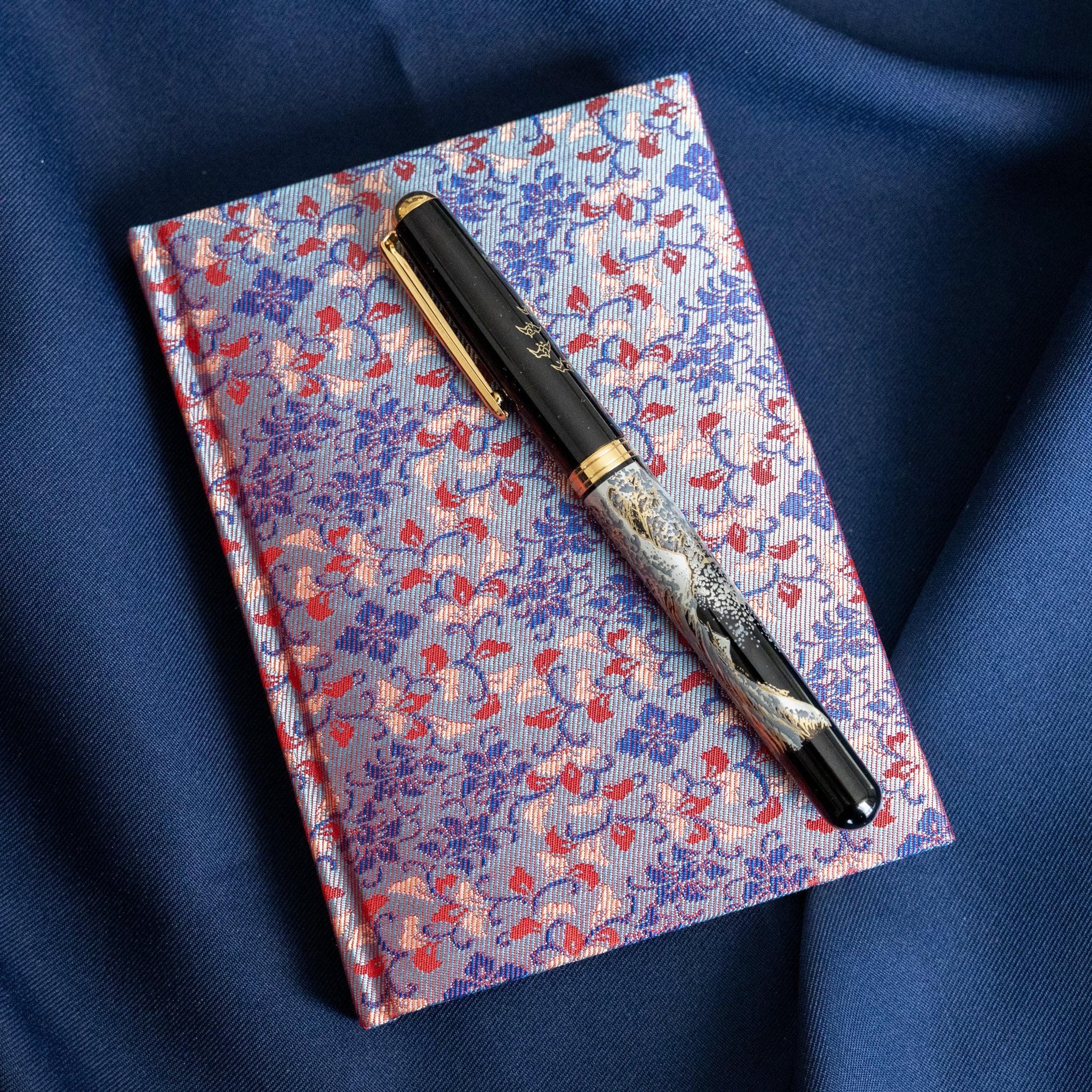 Yusoku Hanabishi Nishijin silk notebook angled with pen on top, lavender-pink and red woven floral cover, blue fabric