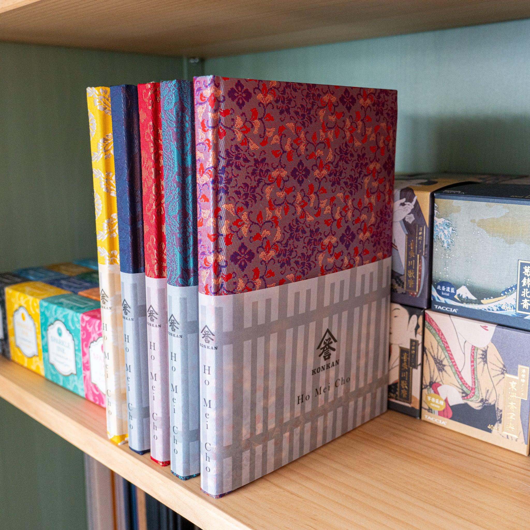 Multiple Yusoku Hanabishi Nishijin silk notebooks on wooden shelf in various colors alongside Japanese gift packaging