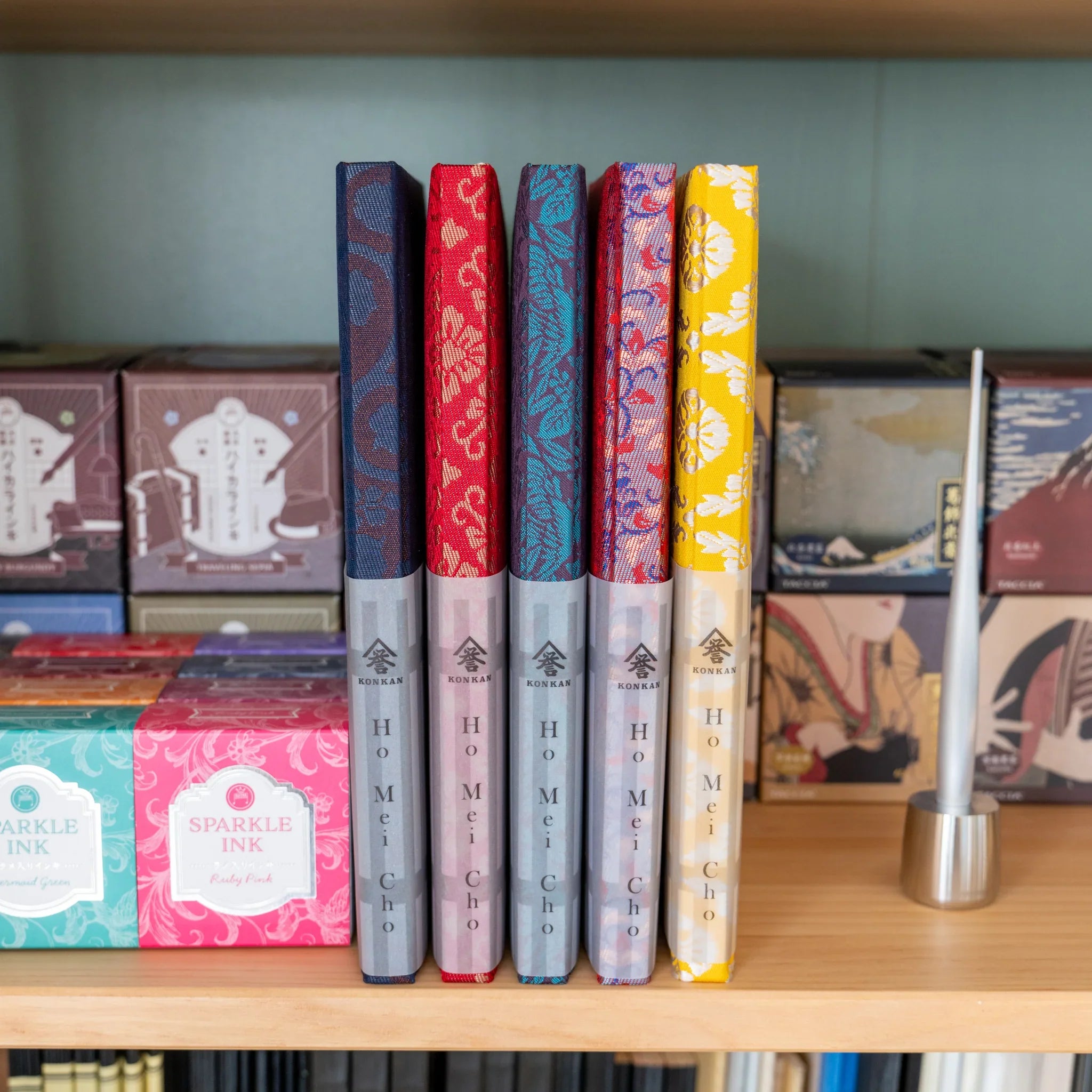 Yusoku Hanabishi Nishijin silk Homeicho notebooks in multiple colors on shelf with stationery items and pen holder