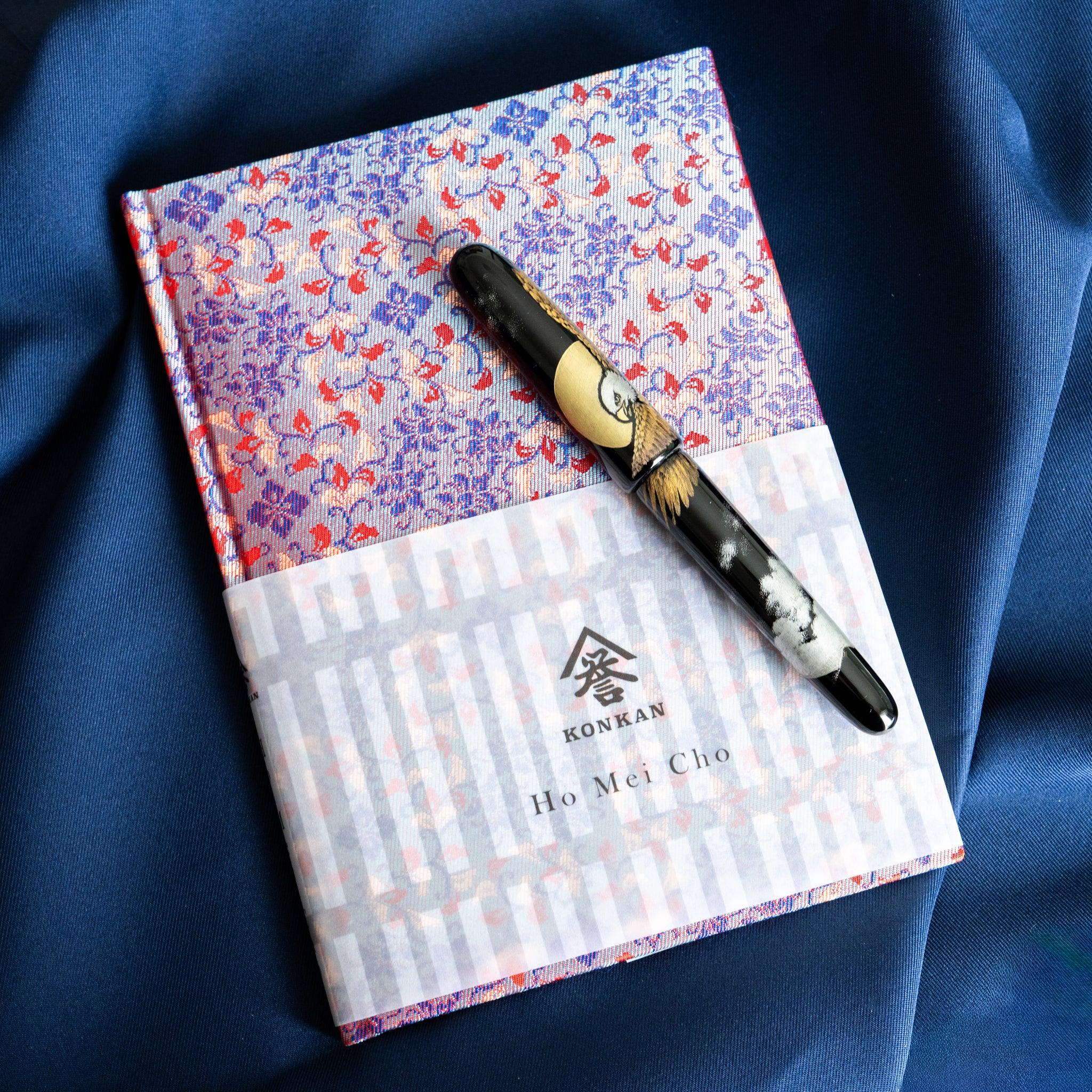 Yusoku Hanabishi Nishijin silk notebook, angled view with pen on top, blue and red woven cover and belly band with KONKAN