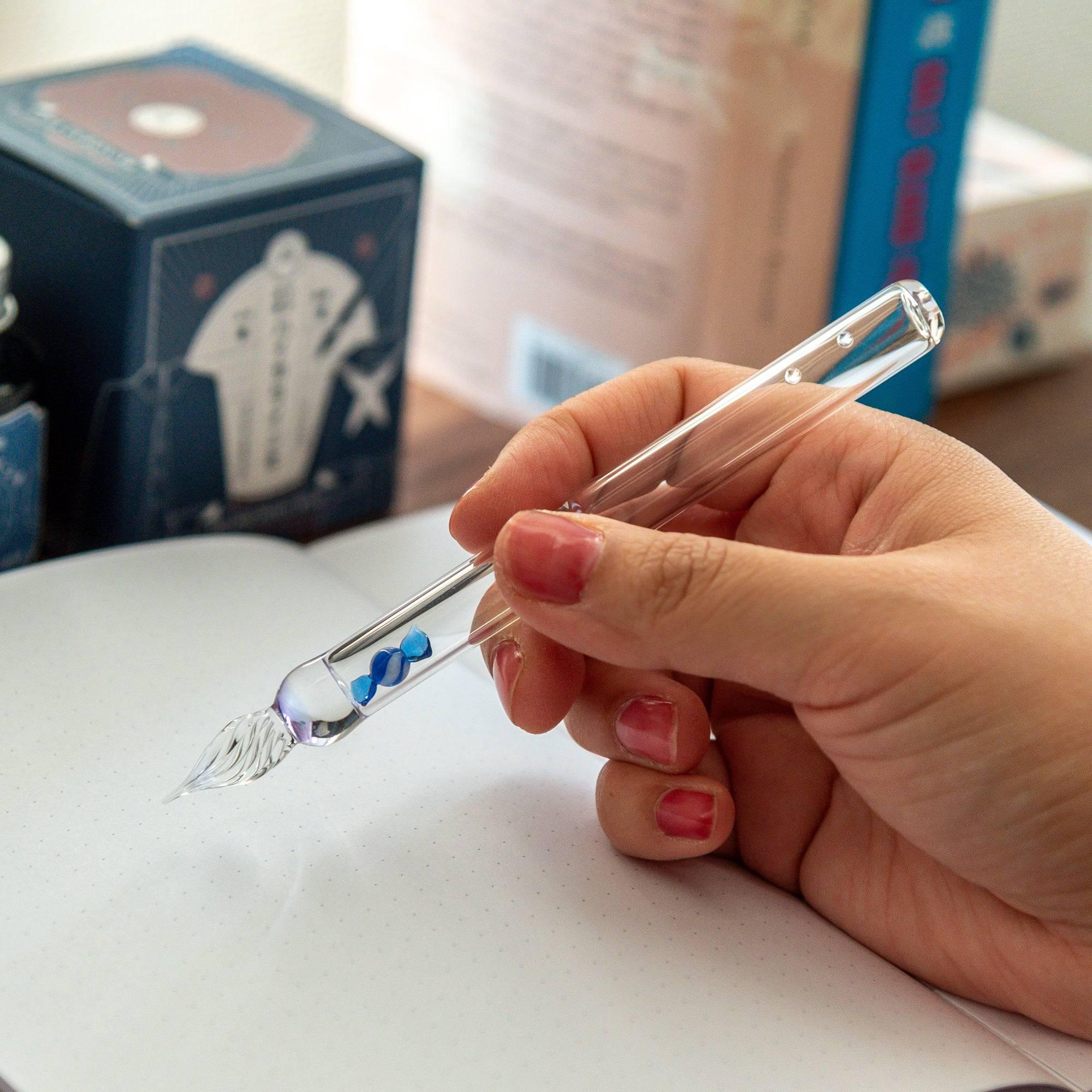 A clear glass dip pen with a blue candy motif encased inside, shown being held while writing, perfect for adding a playful touch to any desk.