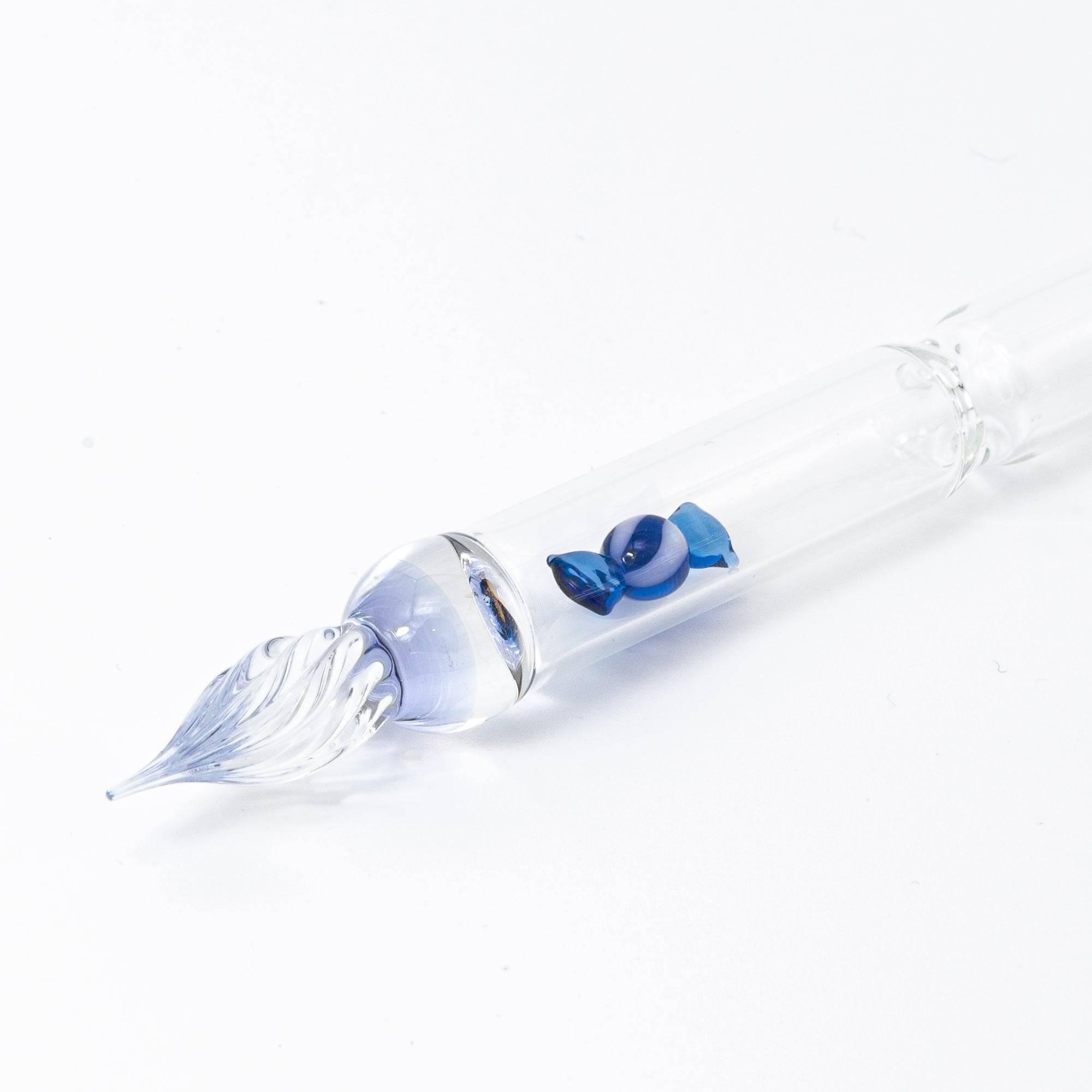 A clear glass dip pen with a small blue candy-shaped glass motif captured inside its shaft. The pen’s nib is intricately designed, enhancing the overall charm.