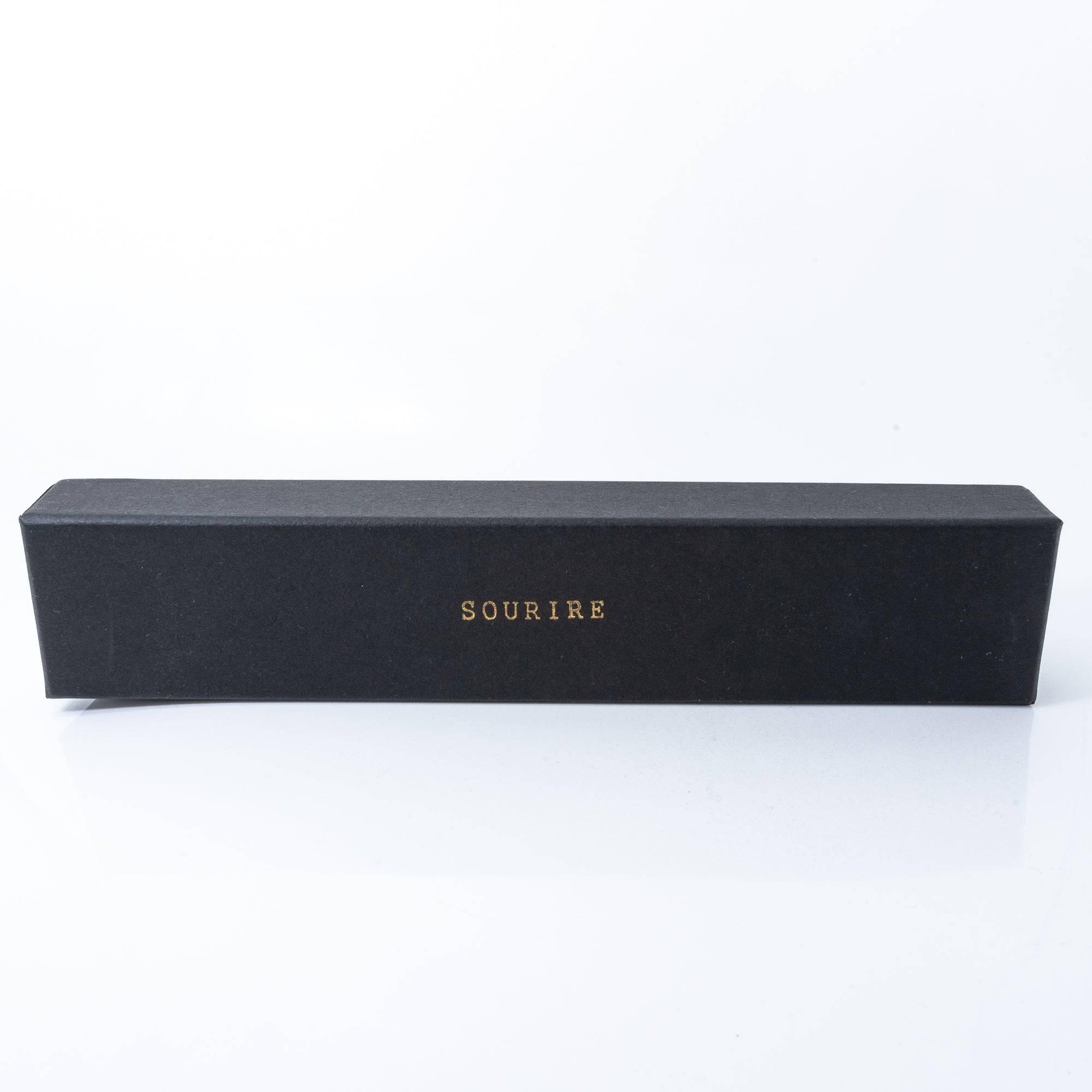 A black box with 'Sourire' written in gold letters on top, containing a beautifully wrapped glass pen.