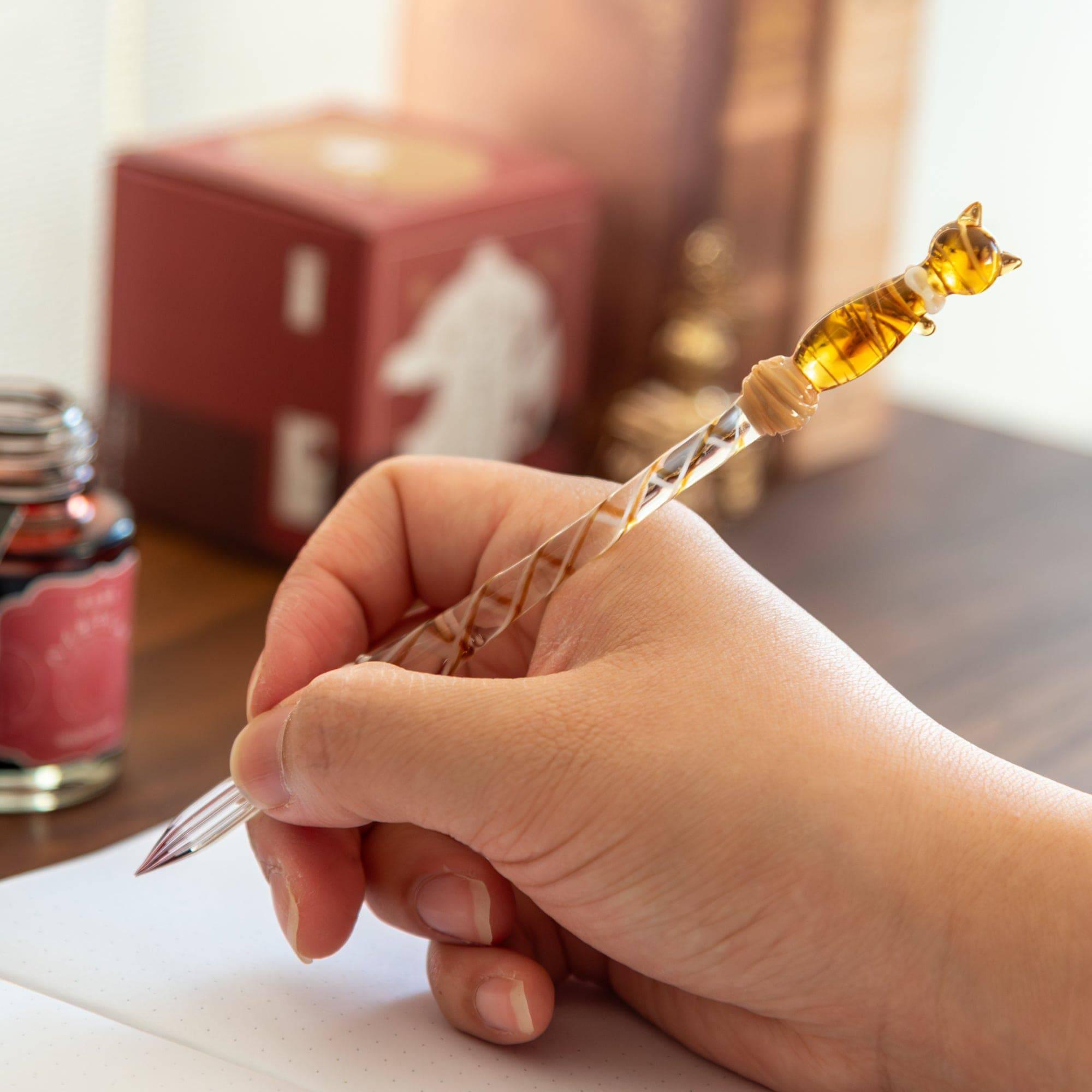 Hand holding a glass pen with a brown cat design, writing in a notebook, showcasing the pen's smooth and elegant use.