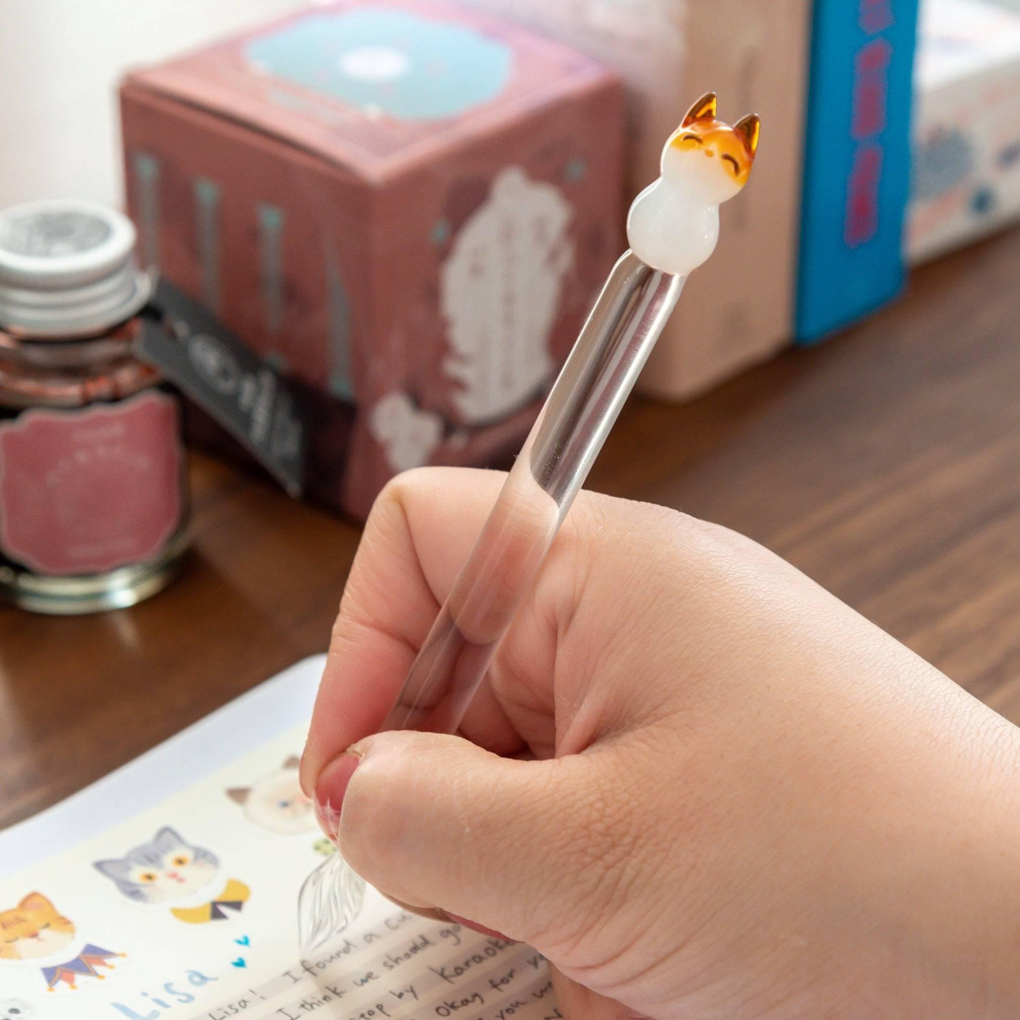 Glass pen with a cat topper and clear shaft, held in hand while writing. Perfect for adding charm to your writing experience.