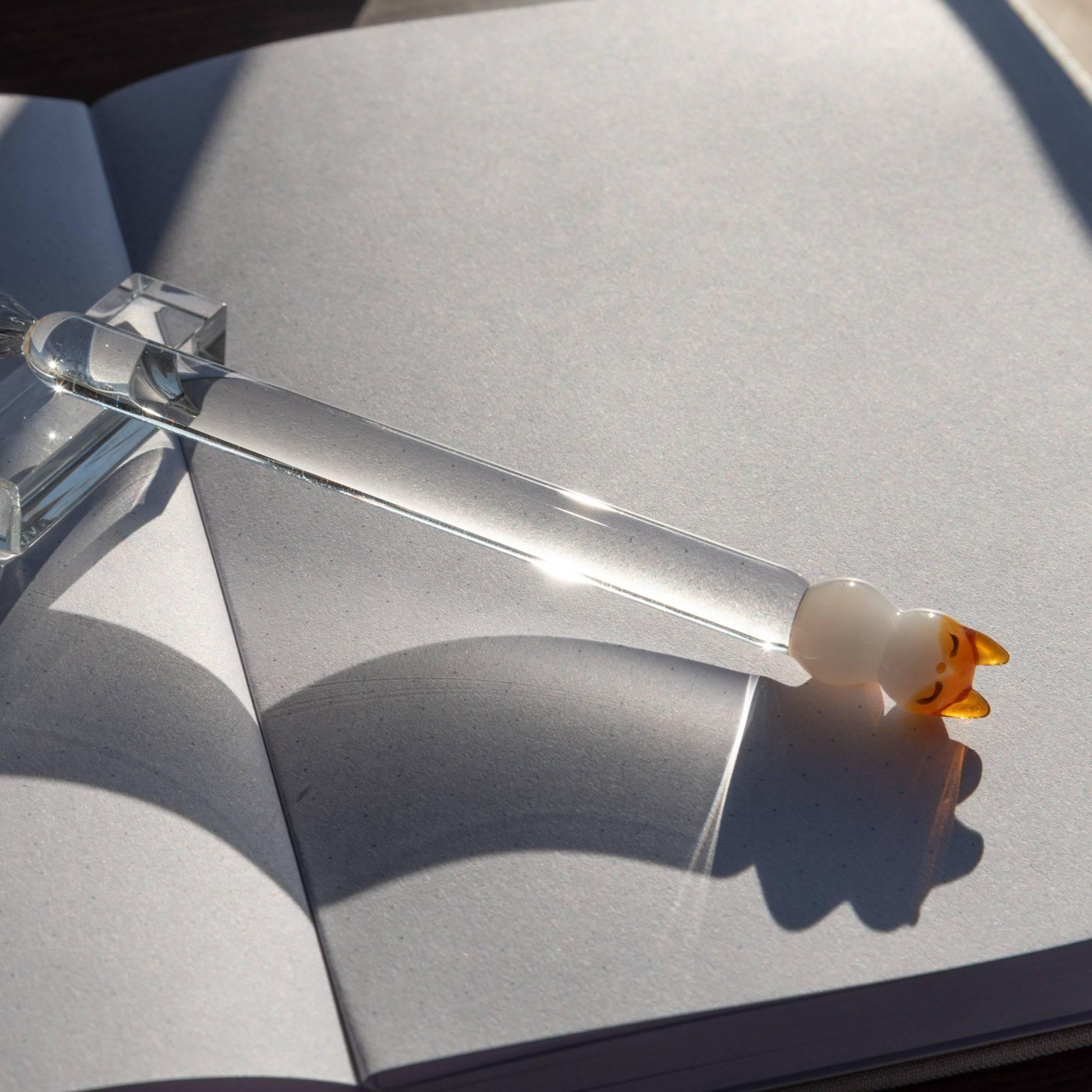 Glass pen with a cat topper and clear shaft placed on an open notebook in sunlight, casting a soft shadow