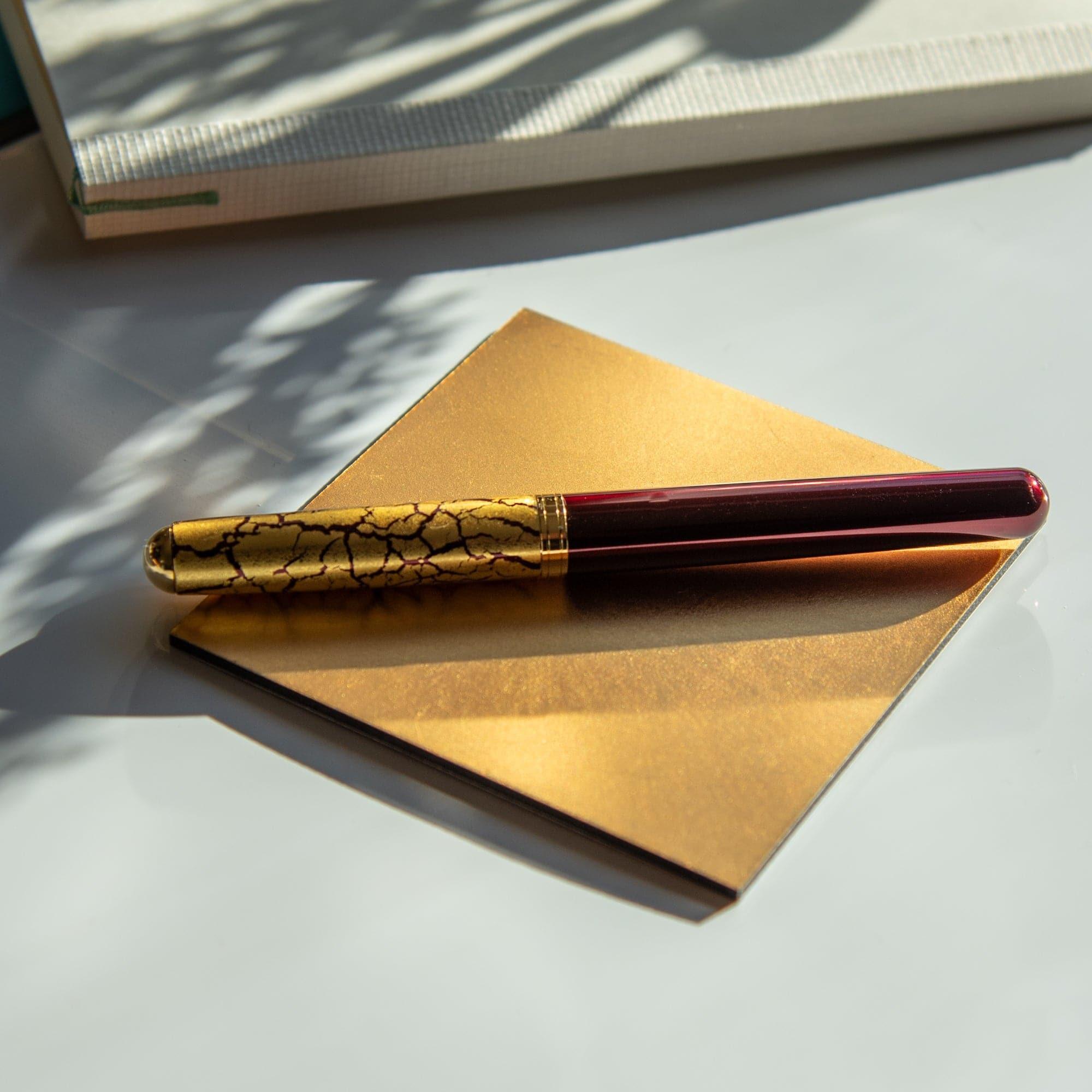 Wine-red fountain pen with a crackled gold-leaf cap displayed diagonally on a golden notepad under soft natural light.