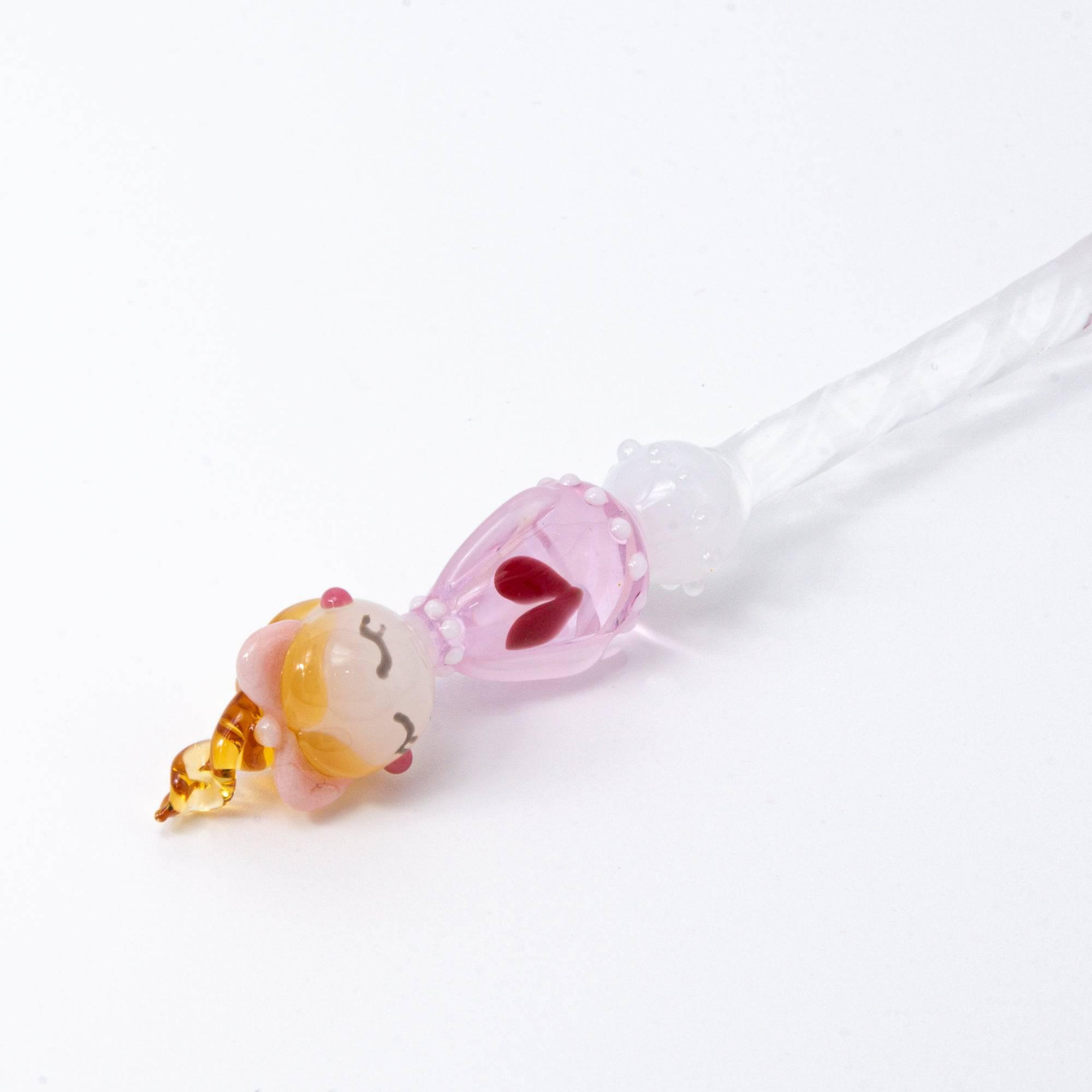 A top view of a Lovery-chan glass pen with a heart design, featuring a cute figure on top, dressed in pink with hearts along the pen shaft.