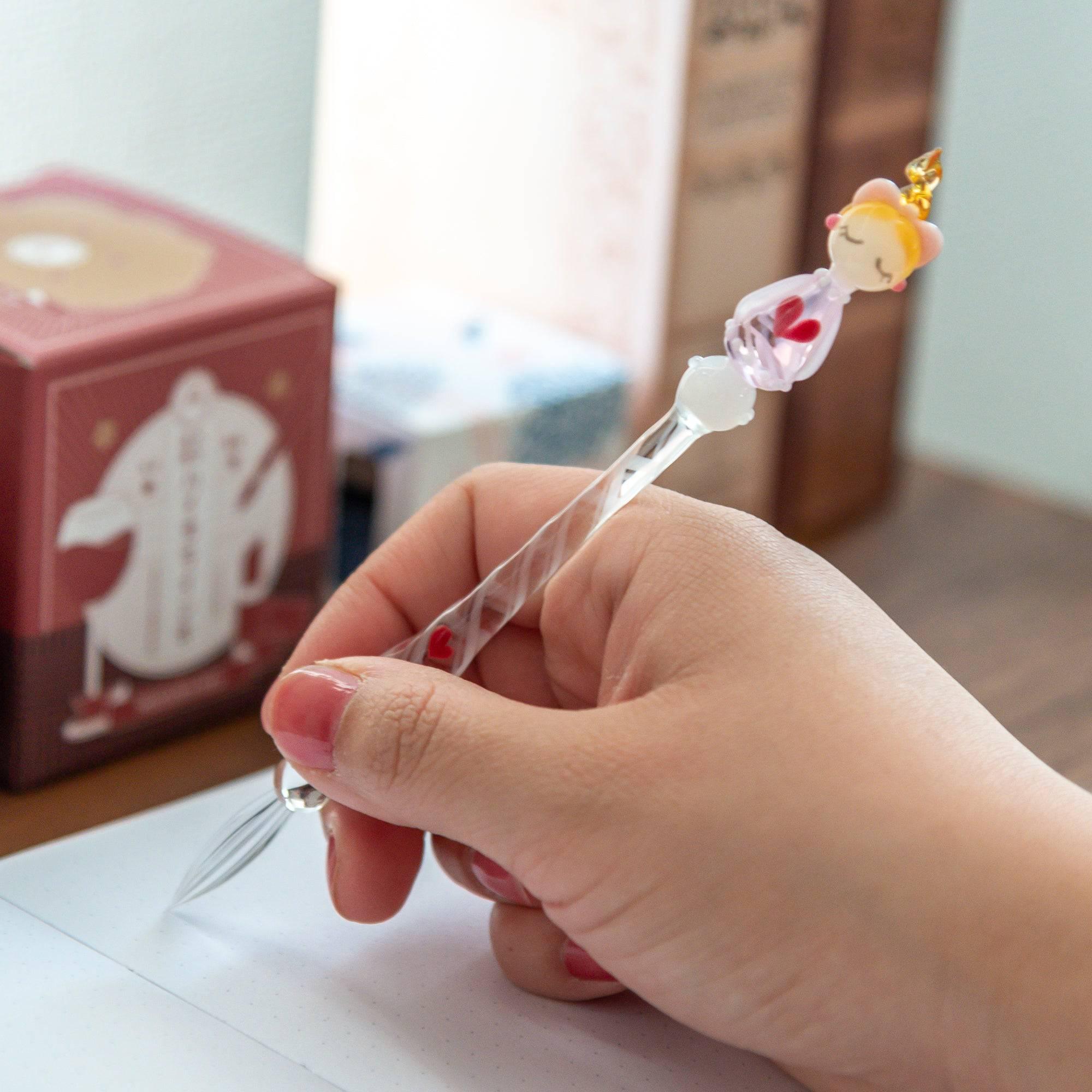 A hand holding a Lovery-chan glass pen with a heart design, featuring a charming pink figure on top, while writing in a notebook.