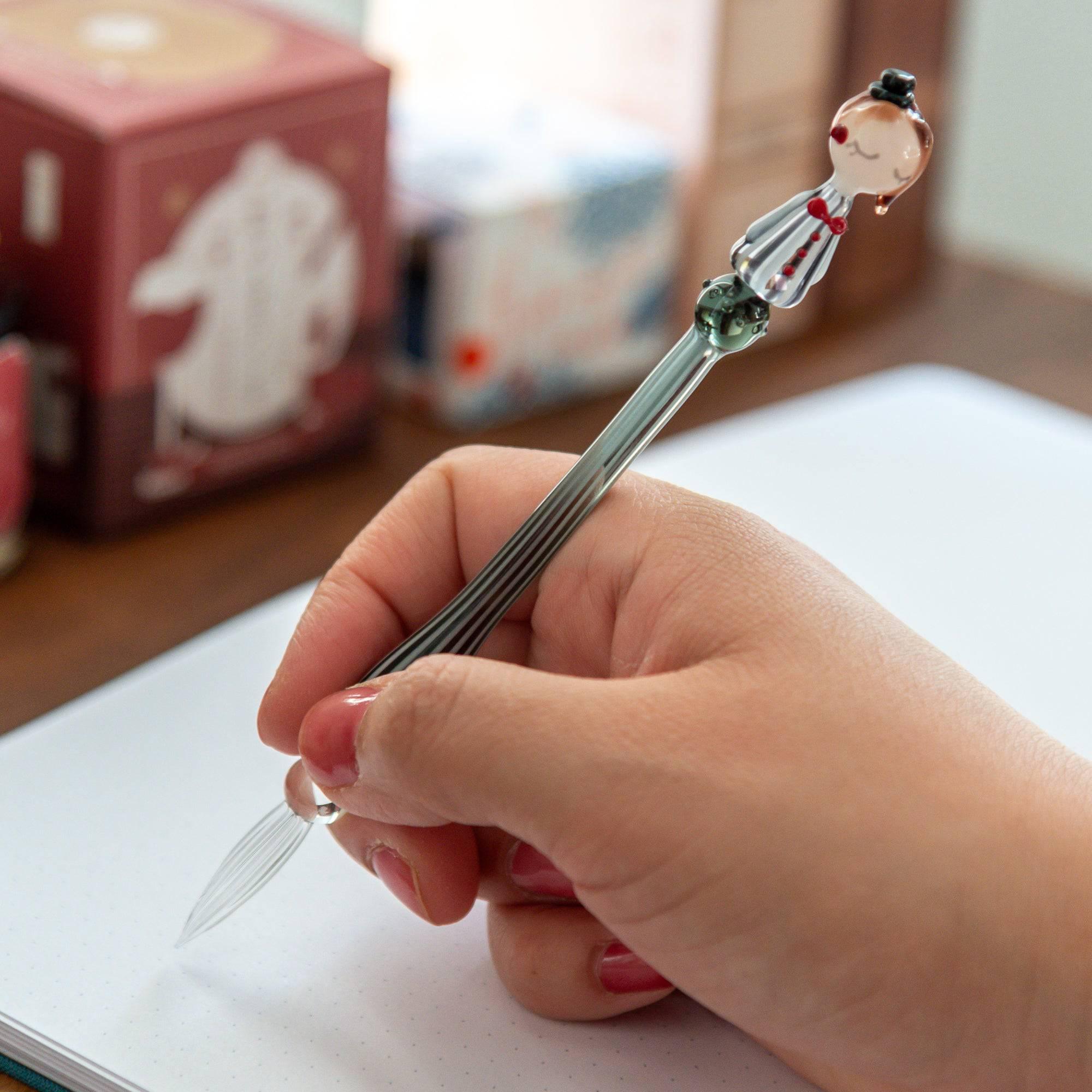 Hand holding a striped glass dip pen with a whimsical lady figurine top, poised for writing in a cozy setting.