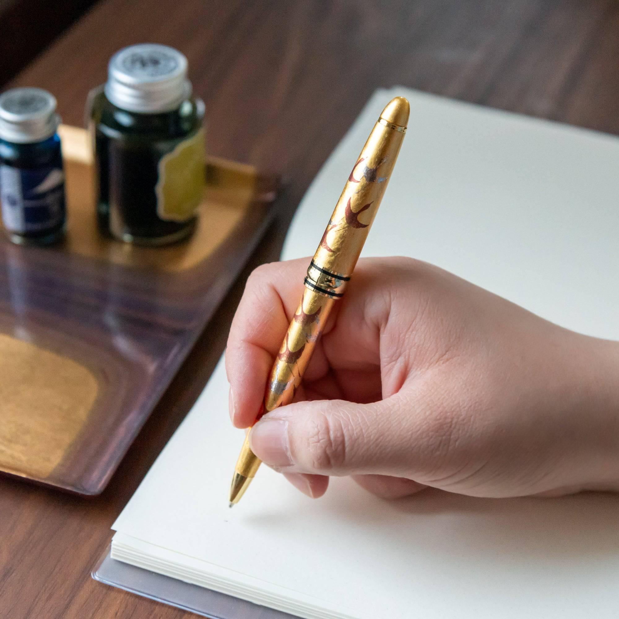 Hand holding the gold maki-e ballpoint pen, poised over a blank notebook, with ink bottles visible in the background on a wooden desk.