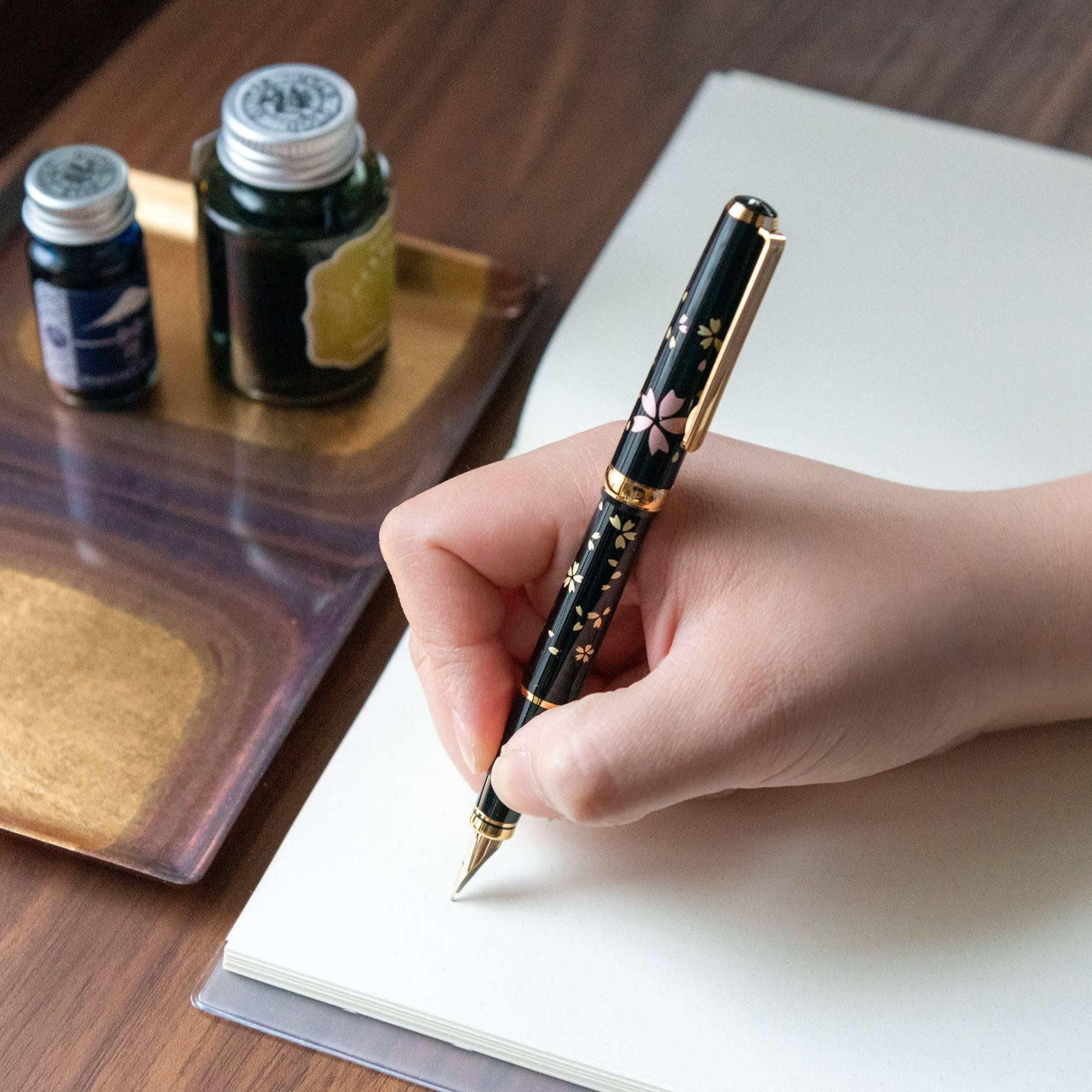 Hand holding a Japanese fountain pen with a black lacquer body, adorned with gold and pink sakura petals, showcasing exquisite craftsmanship.