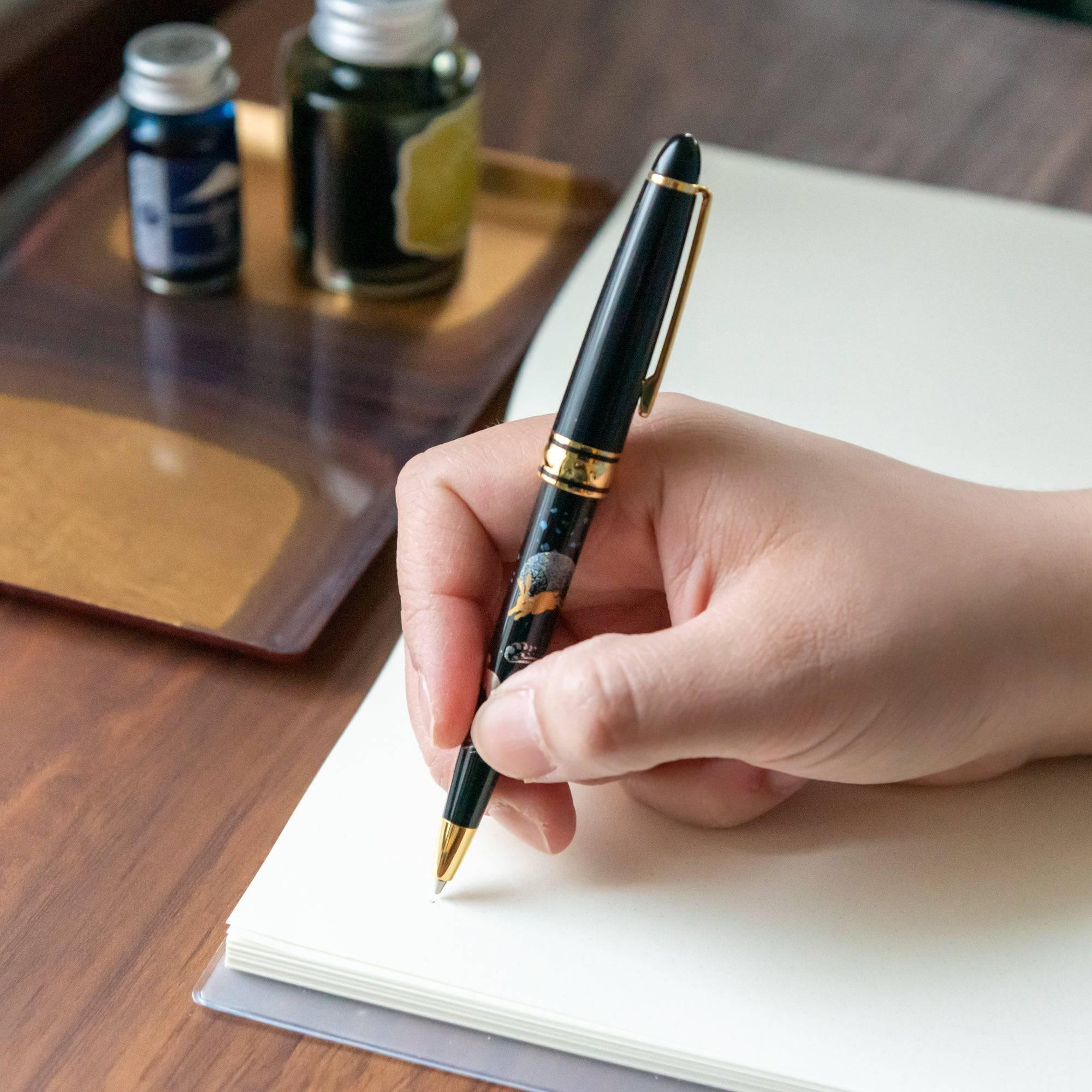 A person writing with a black lacquered pen featuring intricate rabbit and moon designs, with ink bottles on a wooden desk in the background.