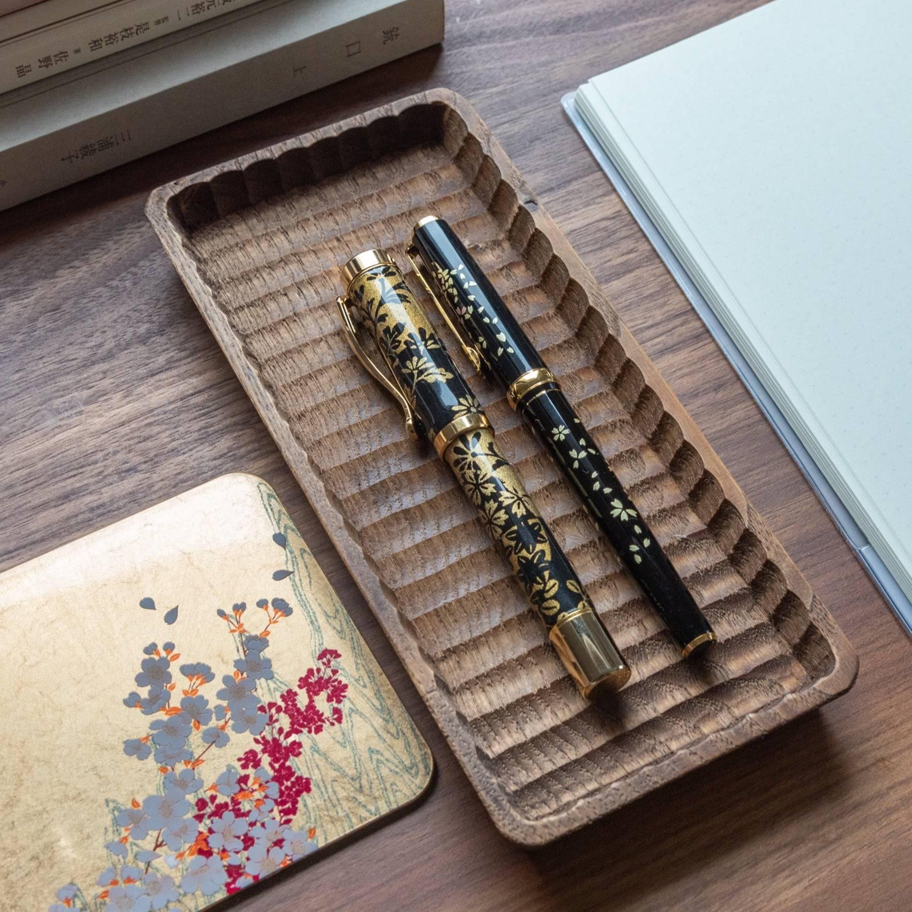 A wooden tray holding two Japanese stationery fountain pens with black and gold floral patterns, placed on a wooden desk with notebooks.  