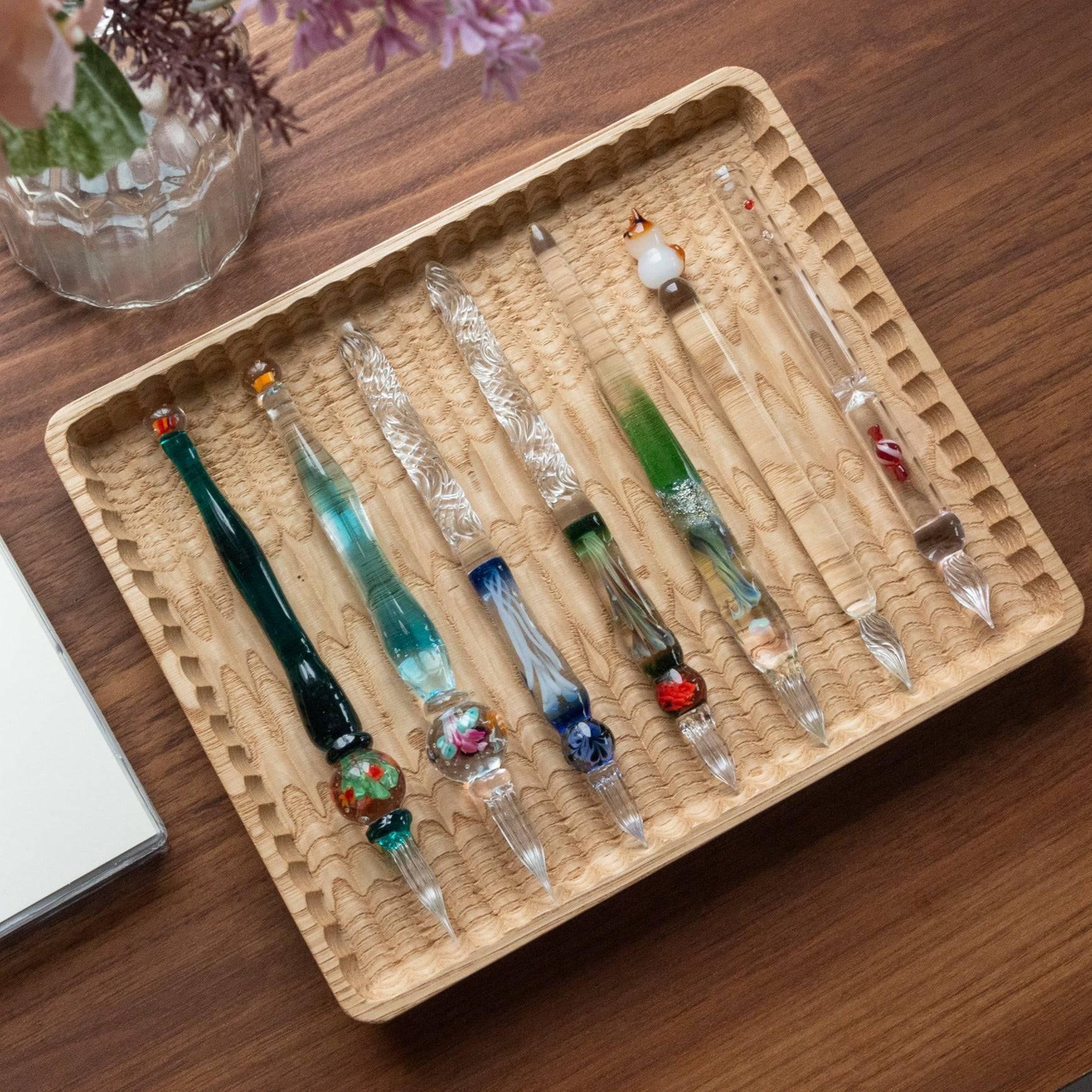 A wooden tray with carved grooves holding an assortment of colorful glass dip pens, placed on a wooden table.  