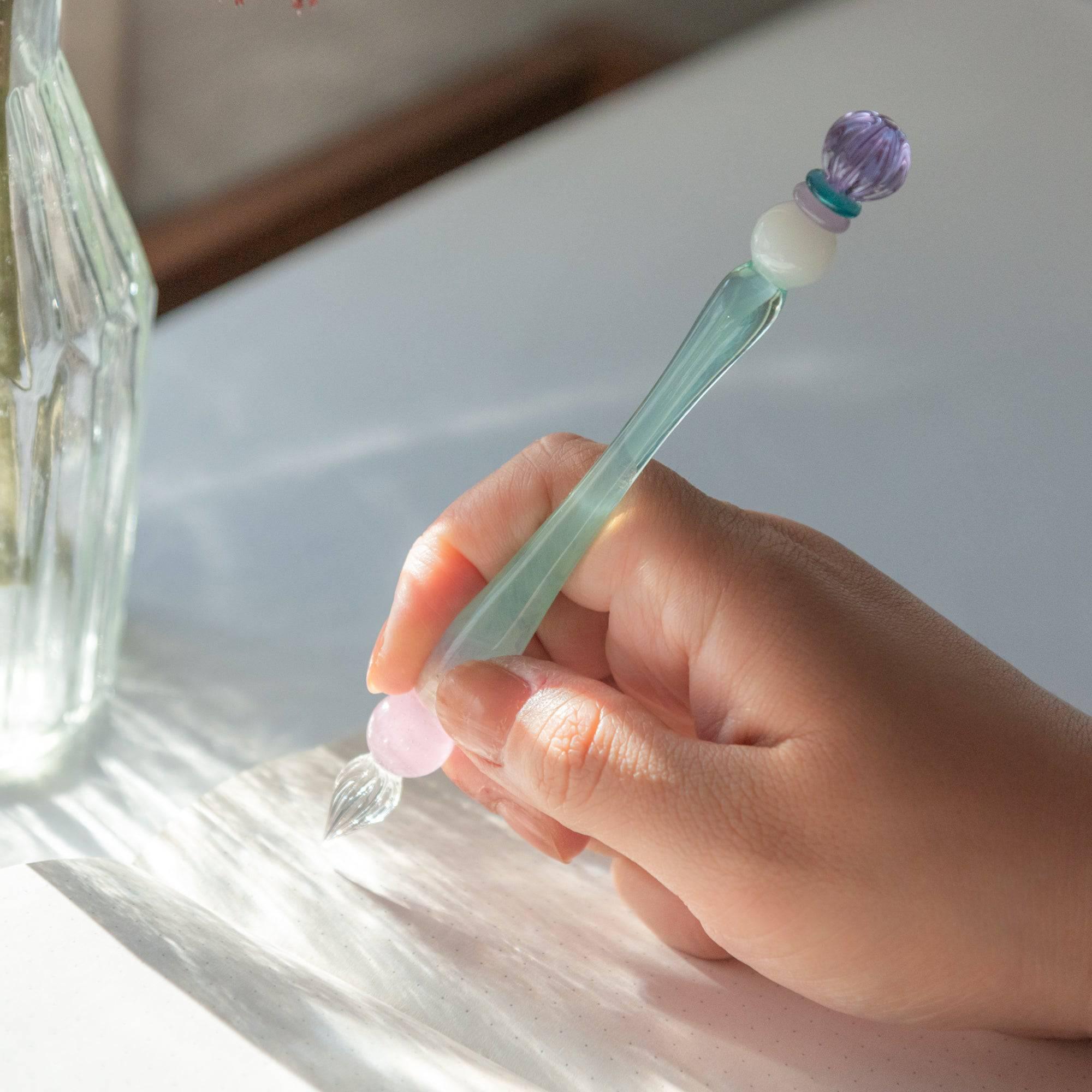 Holding a glass pen with a purple candy-like ornament, blue body, and pink bead, the pen glows as light hits the writing hand.