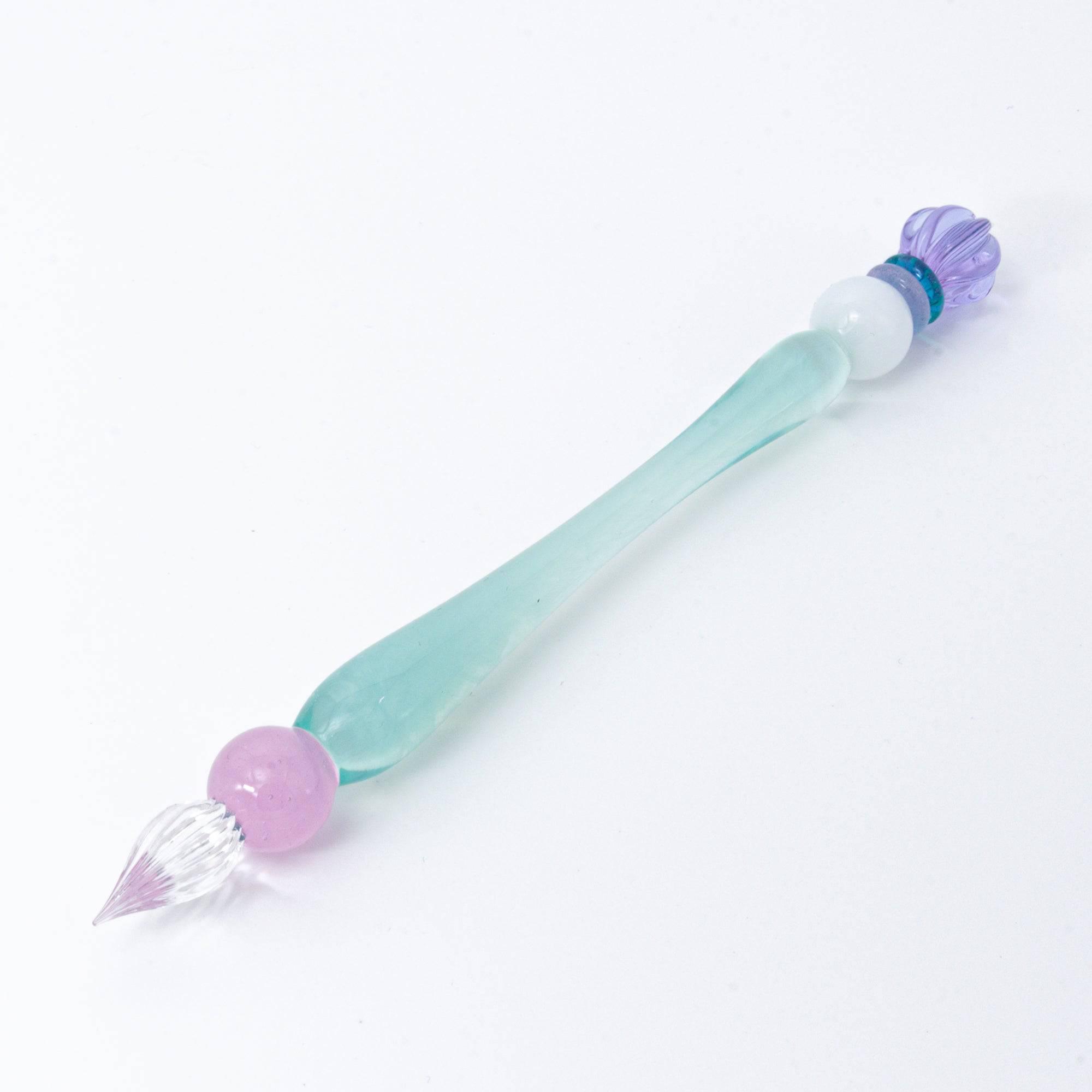 A pastel-colored glass pen with a soft blue body and candy-like top rests on a notebook, bathed in soft sunlight.