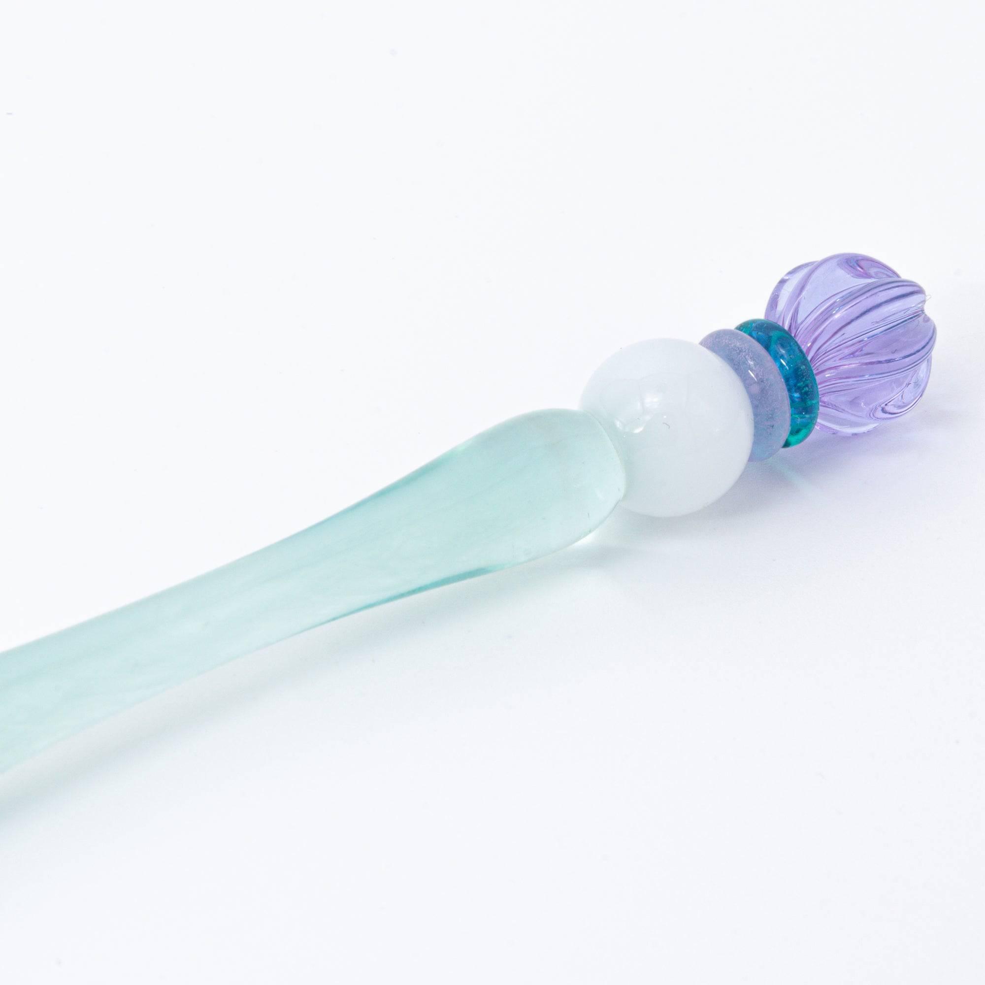 A pastel-colored glass pen with a soft blue body and candy-like top rests on a notebook, bathed in soft sunlight.