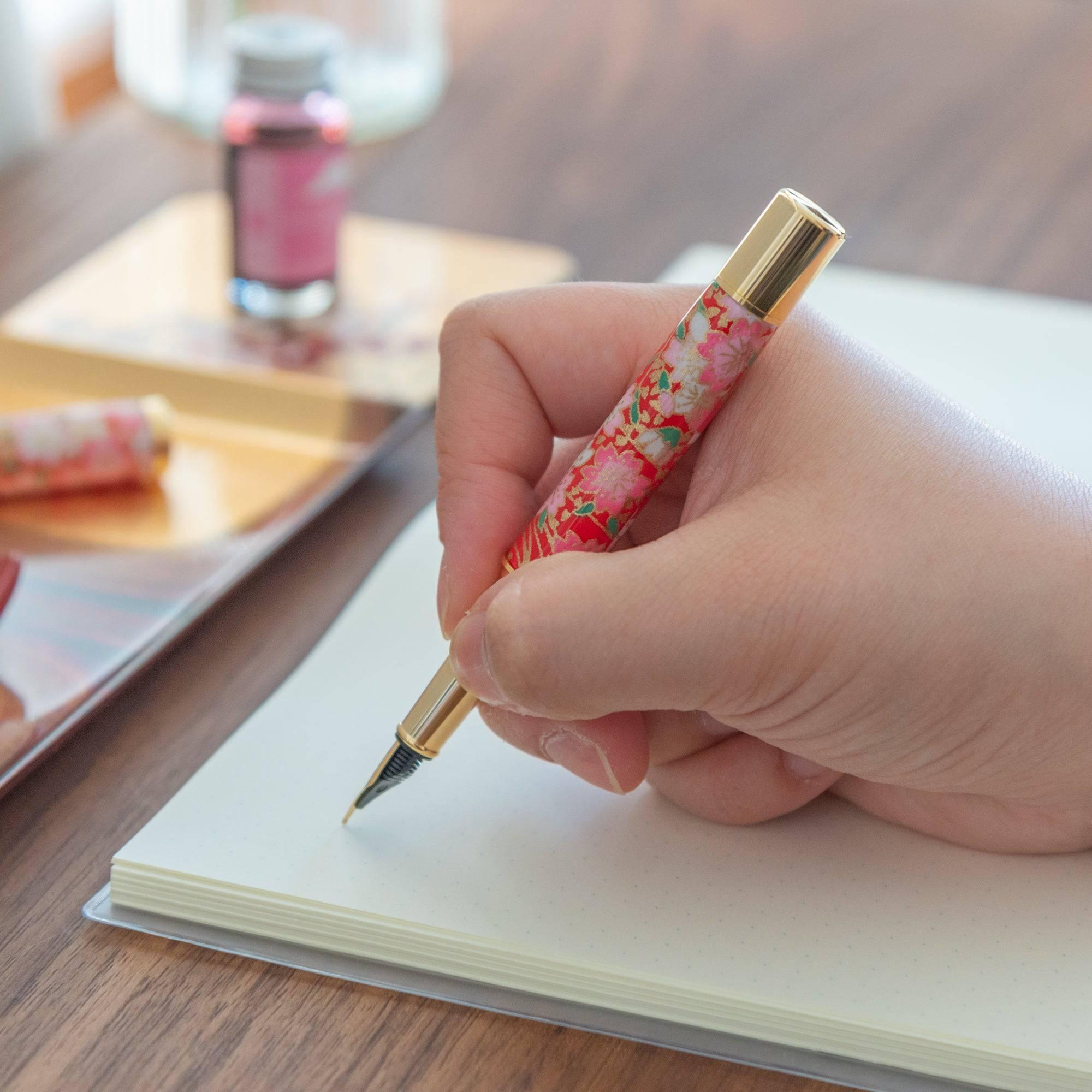 A Japanese fountain pen with a red floral barrel is held while writing, accompanied by a matching ink bottle and pen case in the background.  