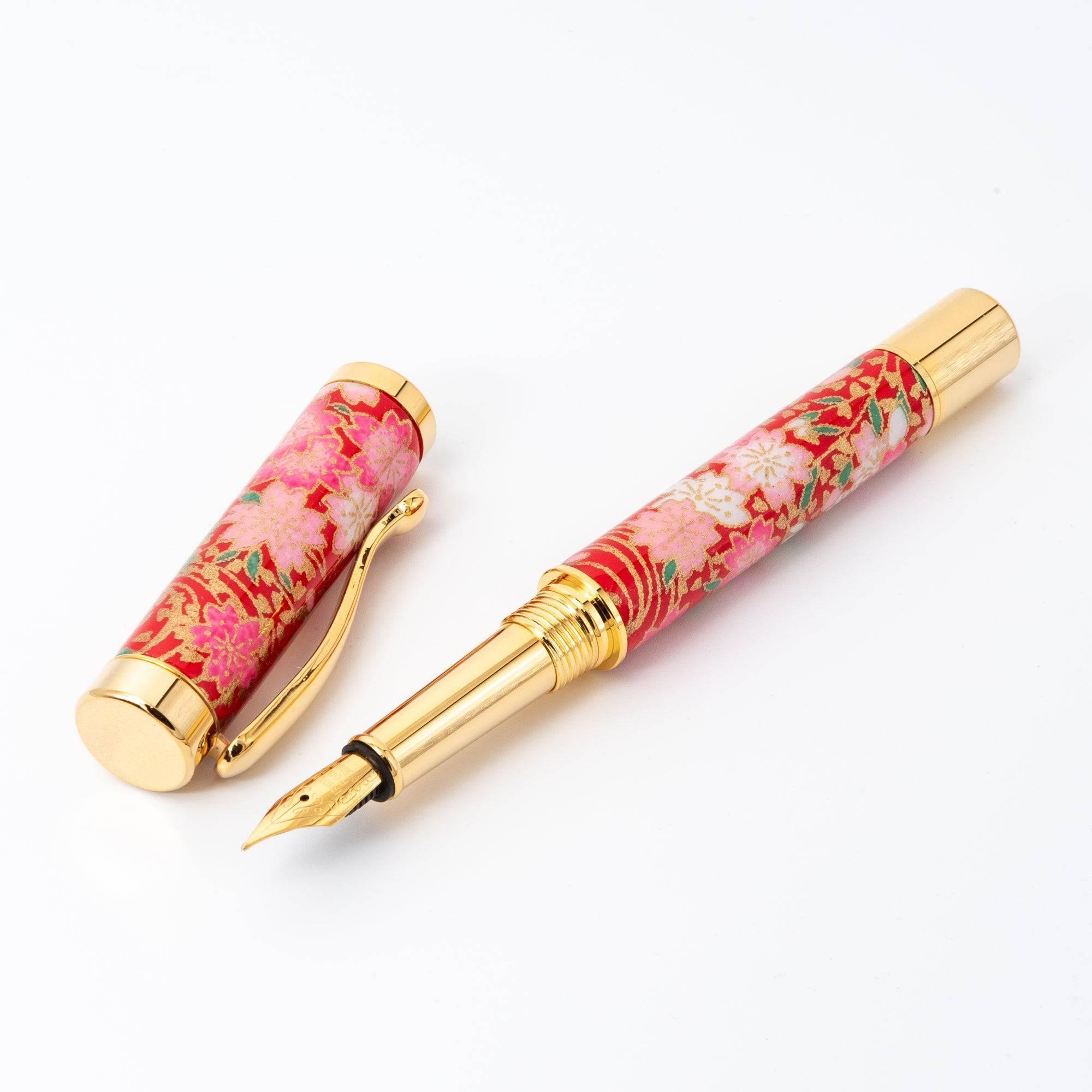 A Japanese fountain pen with a detachable cap, featuring a red floral design and gold details, displayed open to reveal its polished gold nib.  