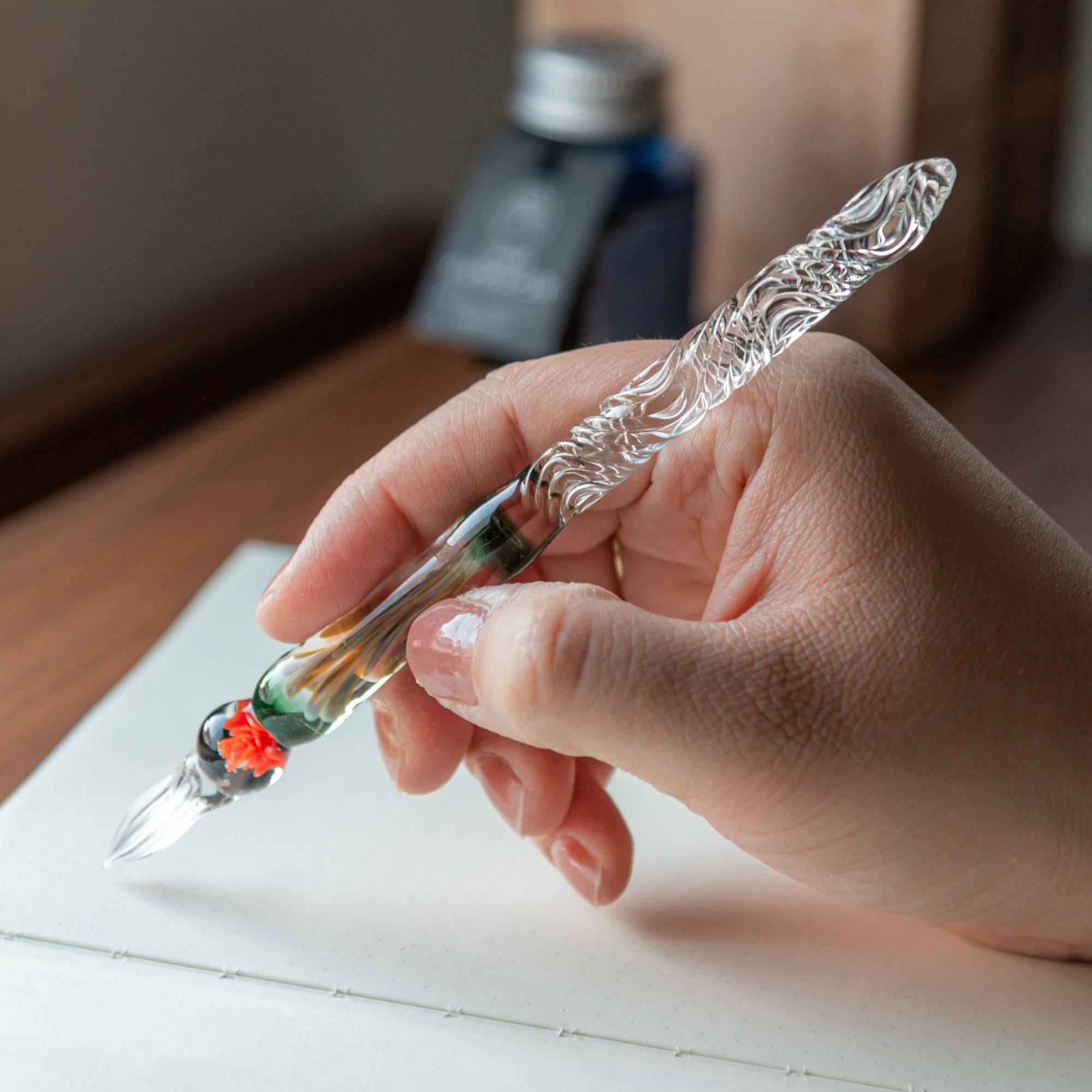 A hand holding a glass pen with an orange rose motif, writing on a notebook, capturing the elegance of the pen in use.