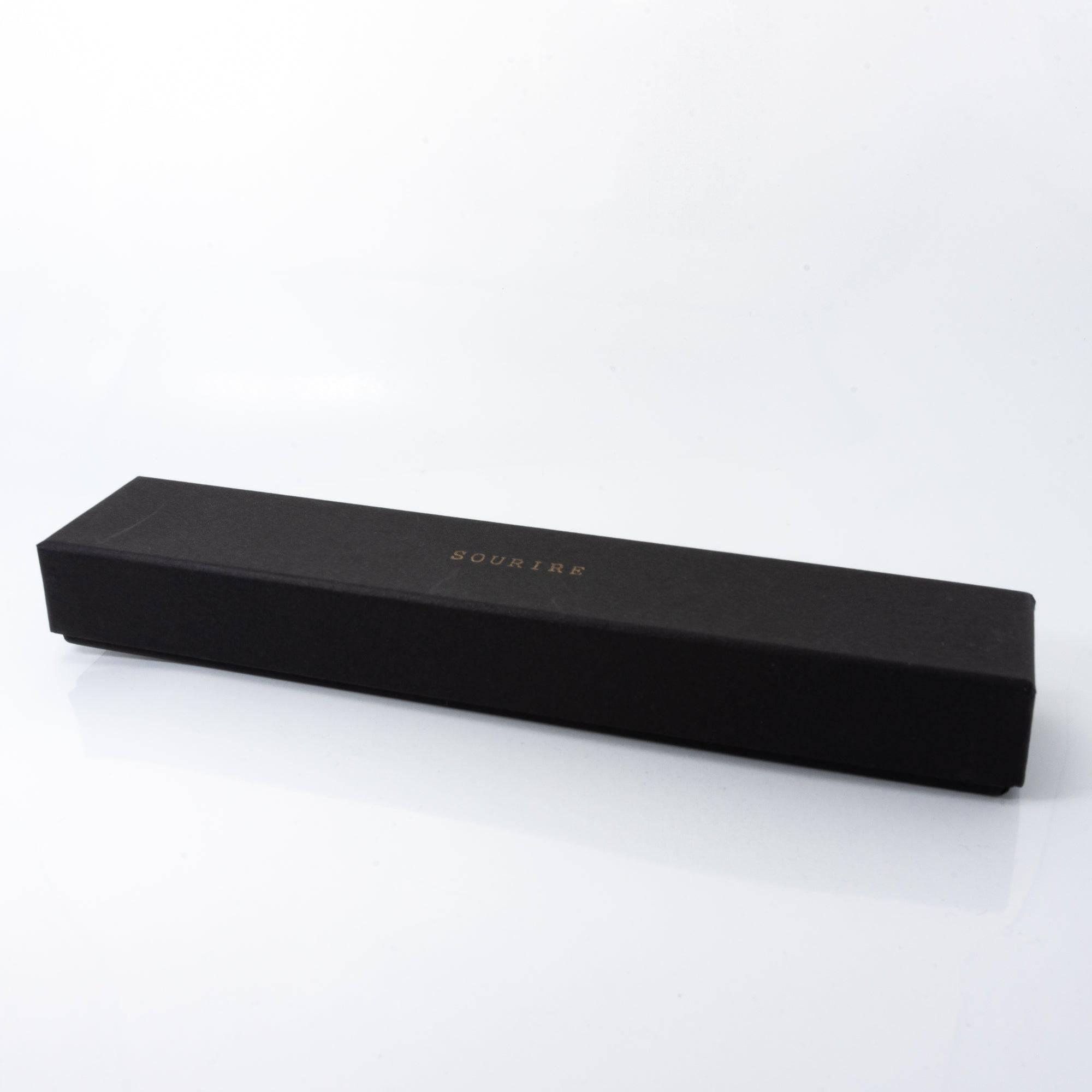 A black box with 'Sourire' written in gold letters on top, containing a beautifully wrapped glass pen.