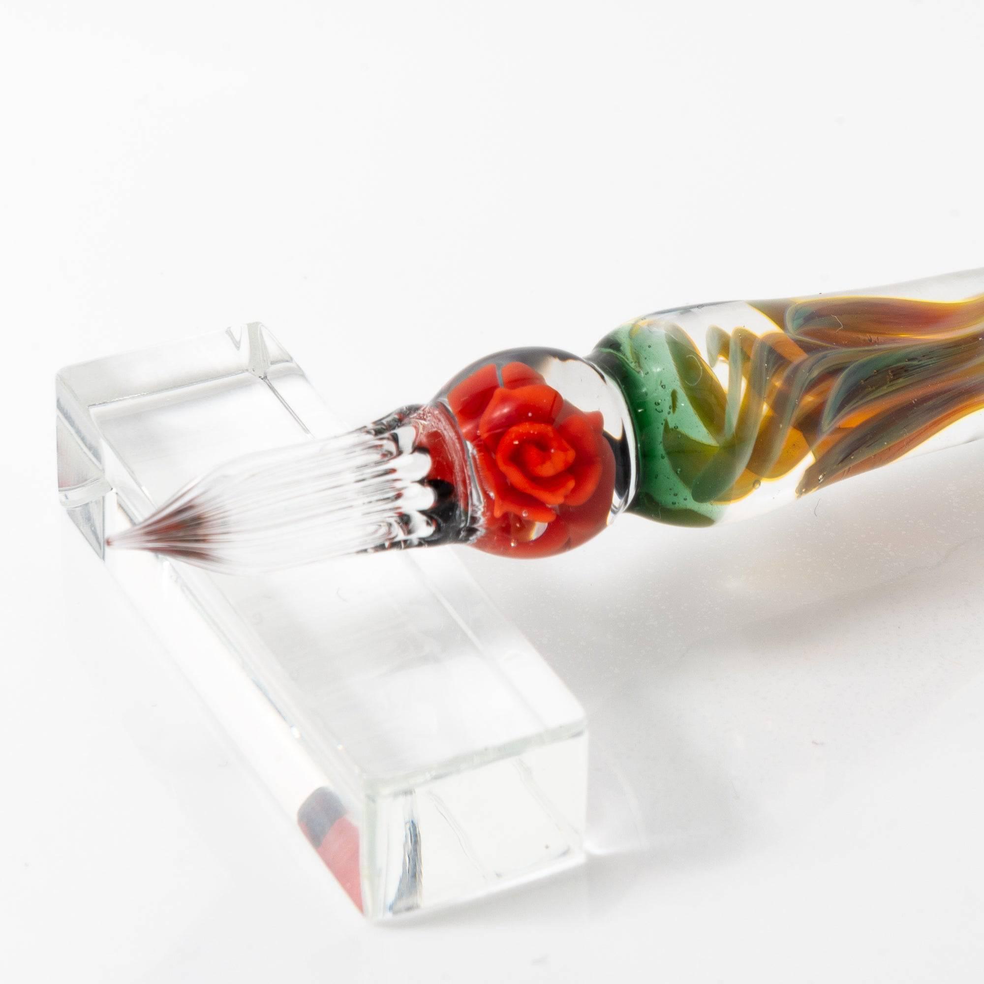 A delicate glass pen with an intricate orange rose motif, resting on a clear pen holder.