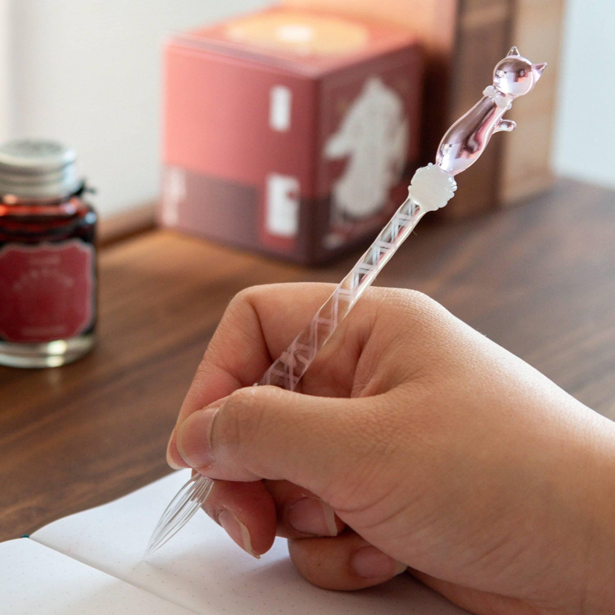 Hand holding a glass dip pen with a pink cat design, writing in a notebook, showcasing the pen's smooth and elegant use.