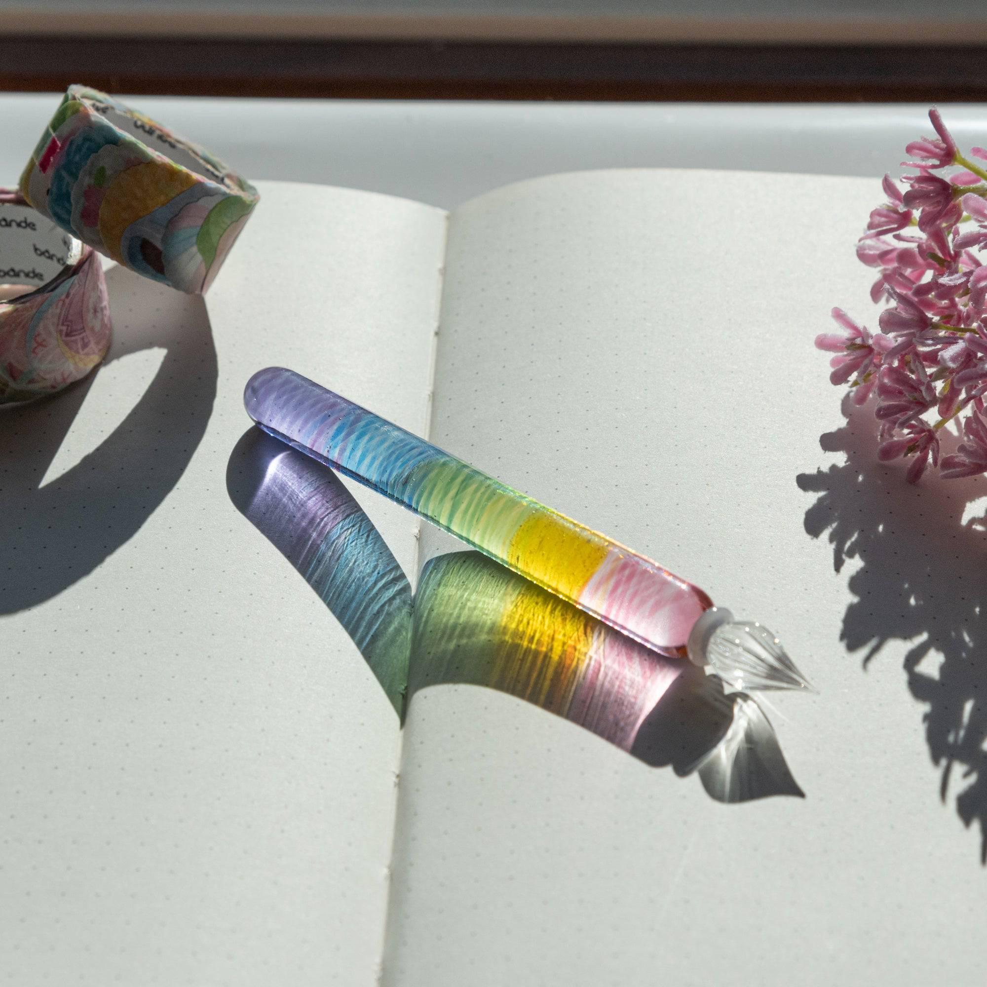 Rainbow-colored glass dip pen reflecting sunlight on an open notebook, accompanied by washi tapes and flowers, ideal for Japanese stationery lovers.