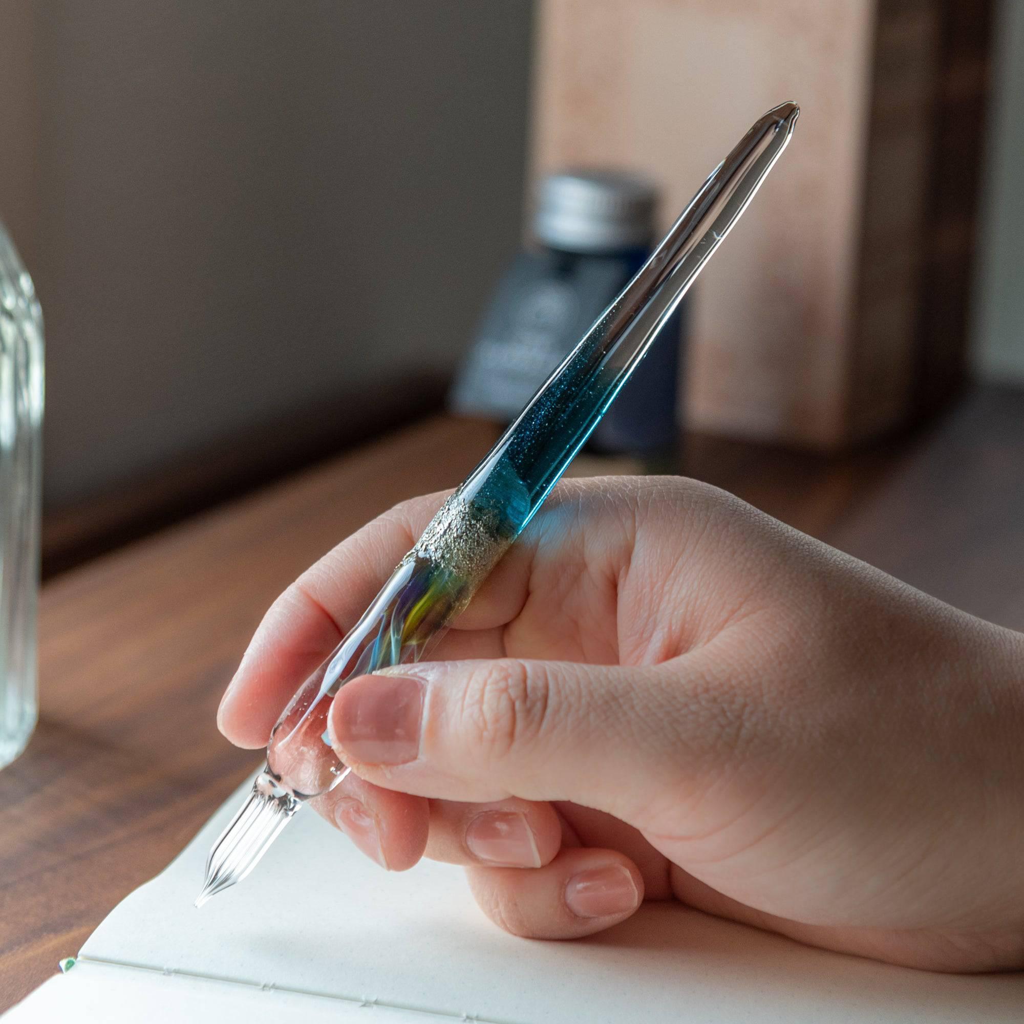 Hand holding a glass pen with a suspended opal and silver accents, featuring a gradient from blue to clear. Ideal for luxury writing experiences.