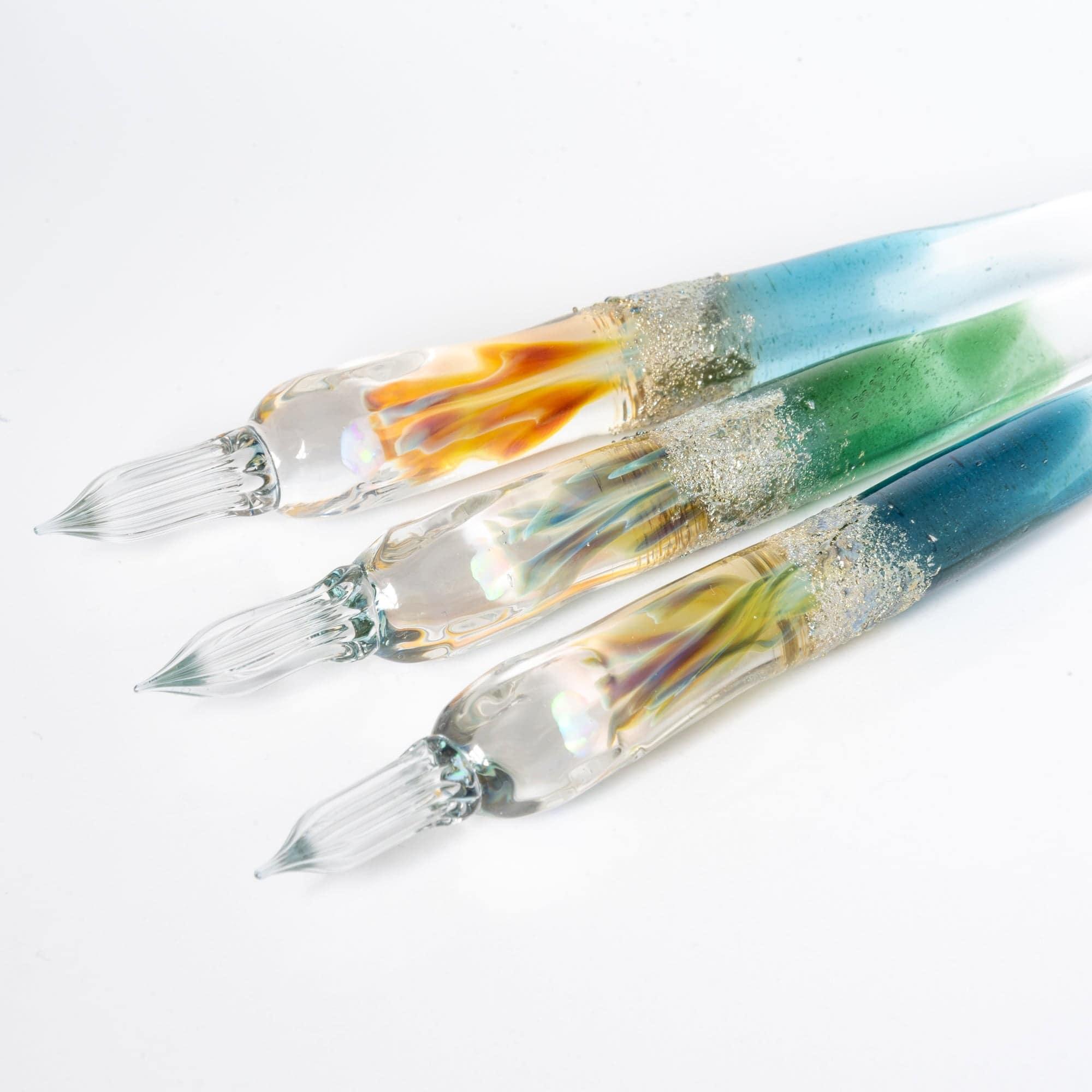 Three glass pens with light blue, green, and clear gradients, suspended opals, and silver accents, displayed in sunlight for a luxurious look.