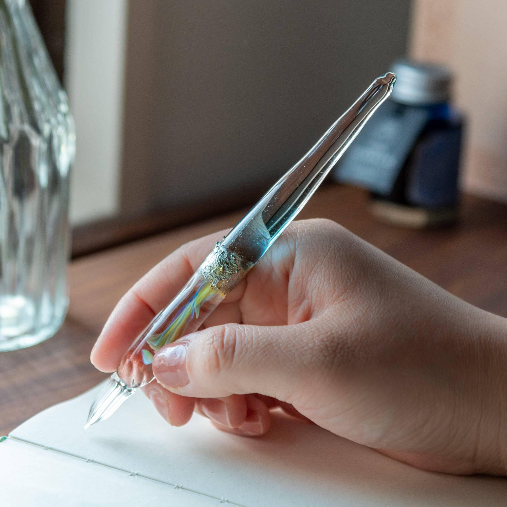 Hand holding a glass pen with a light blue gradient, suspended opal, and silver accents, ideal for elegant writing experiences.