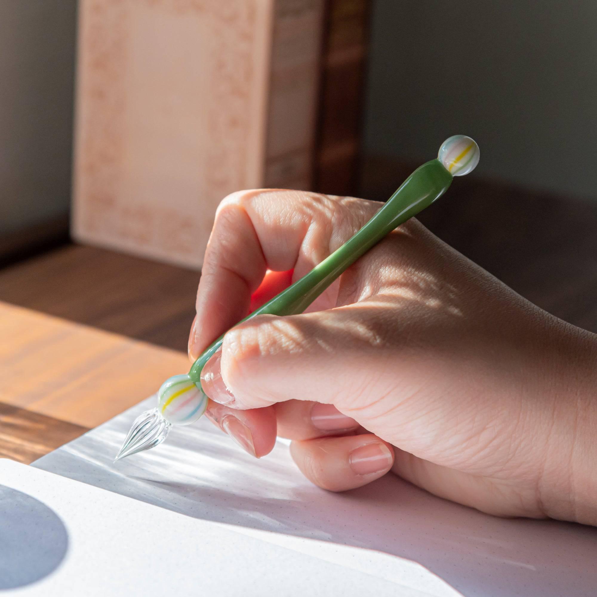 A person writing with a green matcha color glass pen, the sunlight casting soft shadows, highlighting the pen's delicate design and colorful marble details.