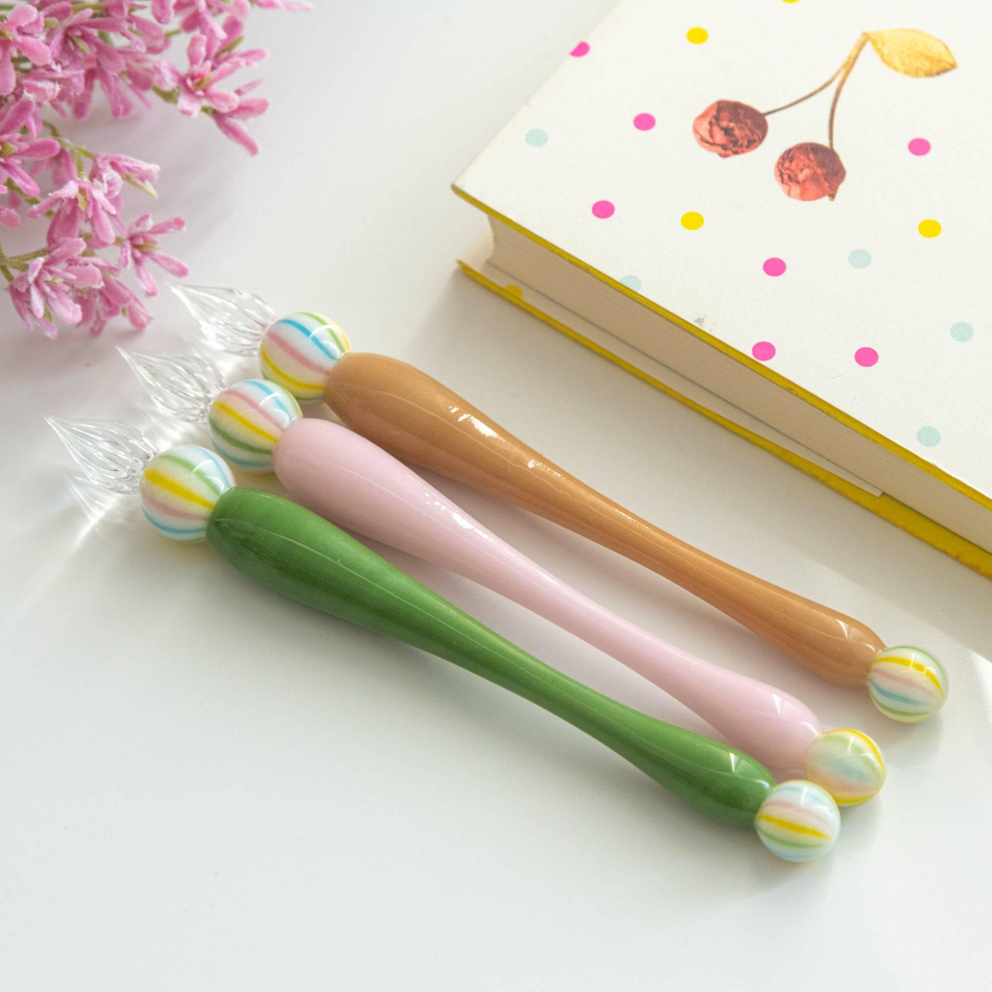 Three glass pens in green, pink, and brown, each with a colorful marble-like accent at both ends, arranged next to a cheerful polka-dotted notebook.