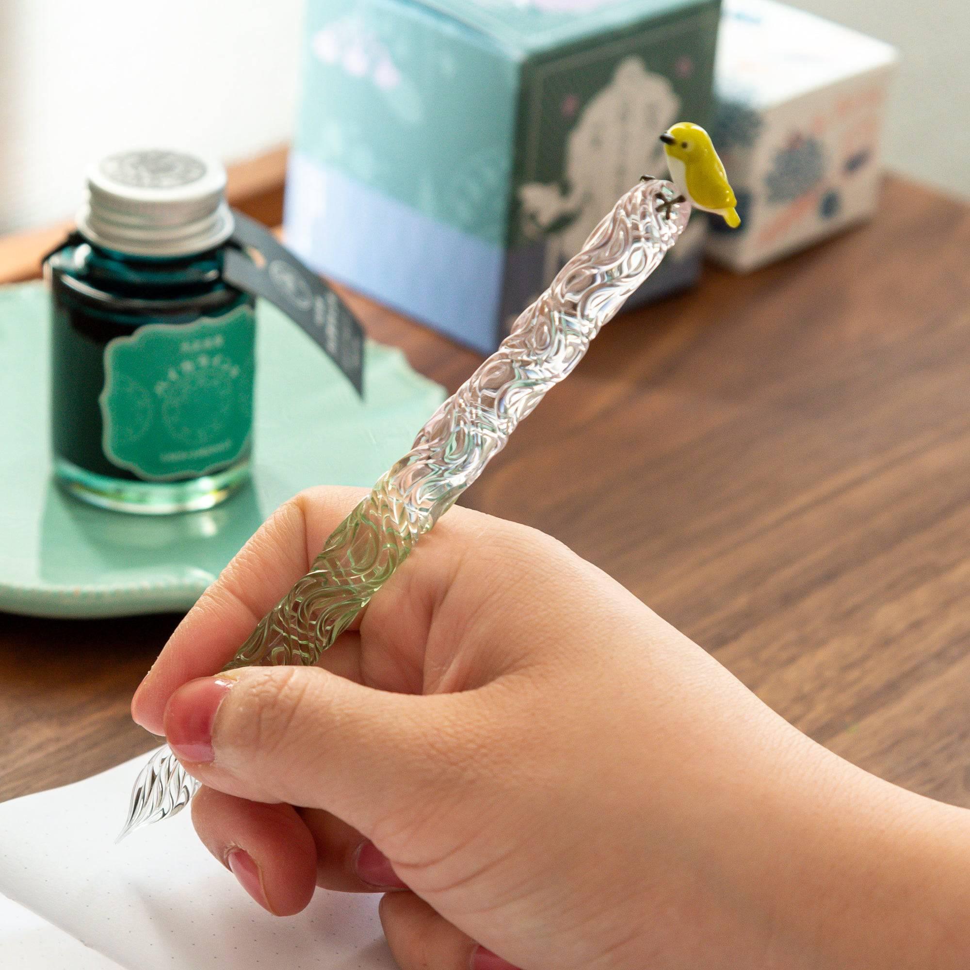 Glass pen with a bird topper and a two-tone twisted shaft in green and pink, held in hand while writing. A charming addition to your stationery.